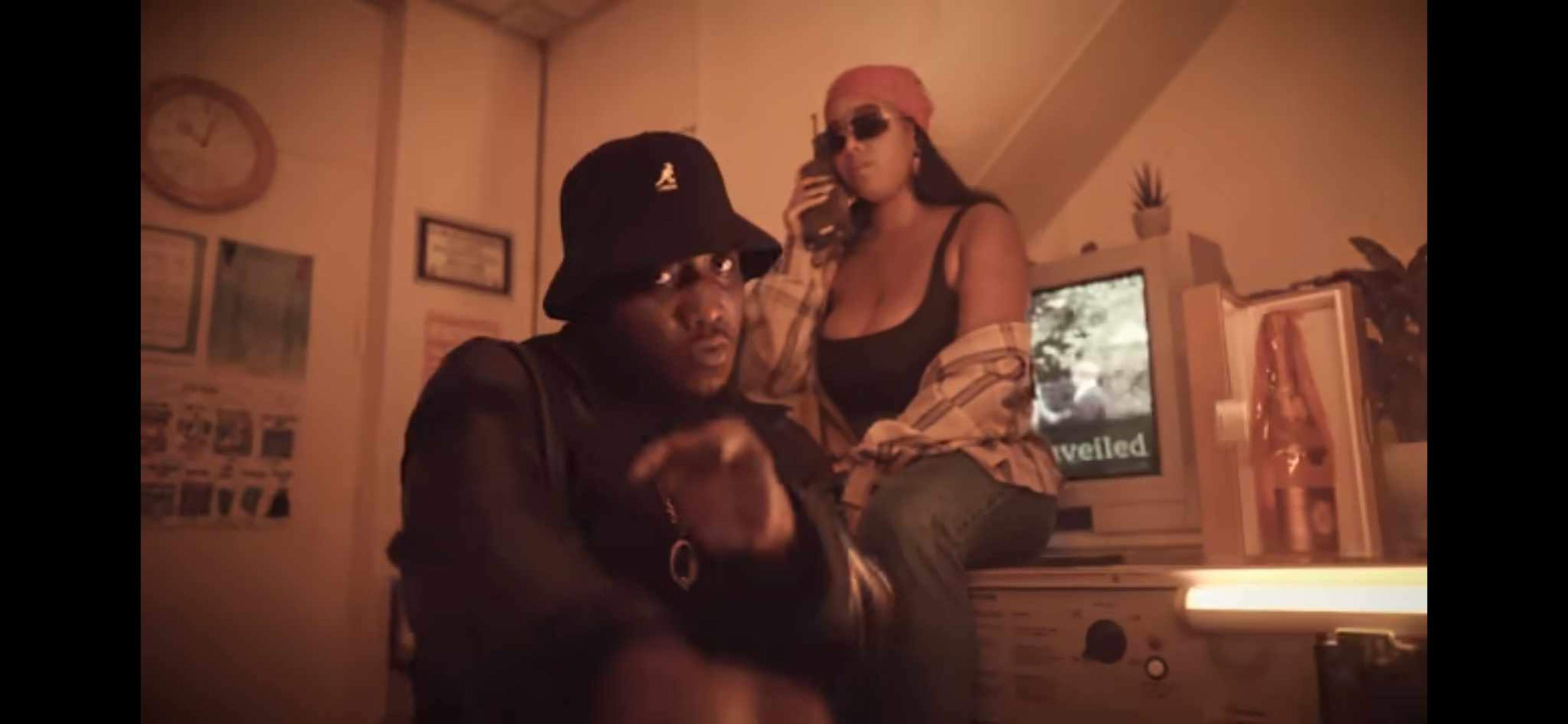 A man in a black bucket hat and sunglasses is seated in front of an old computer, while a woman wearing a pink beanie and sunglasses sits behind him, holding a phone.