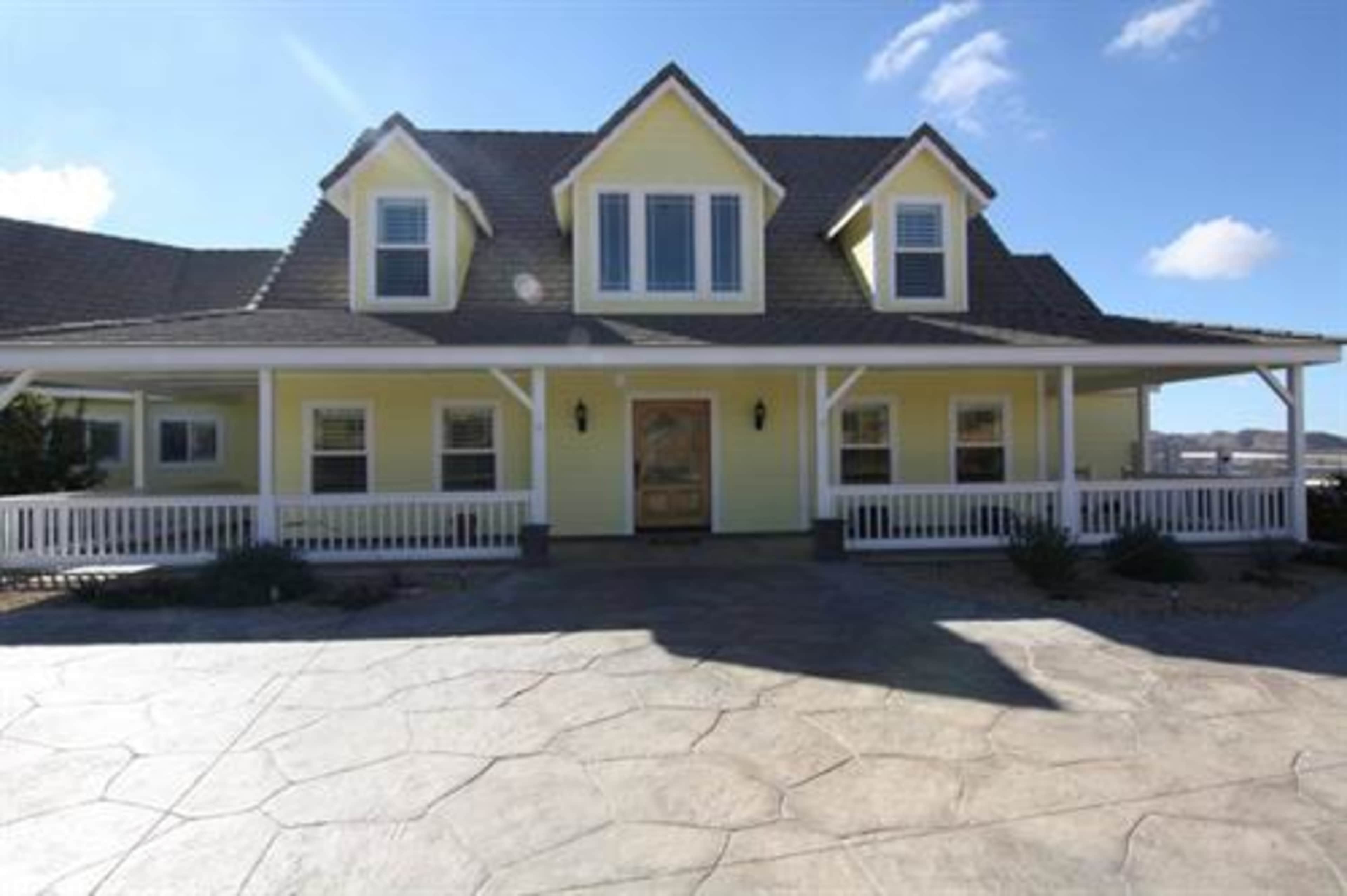 A yellow two-story house with a gabled roof and a front porch sits on a stone patio under a clear sky.