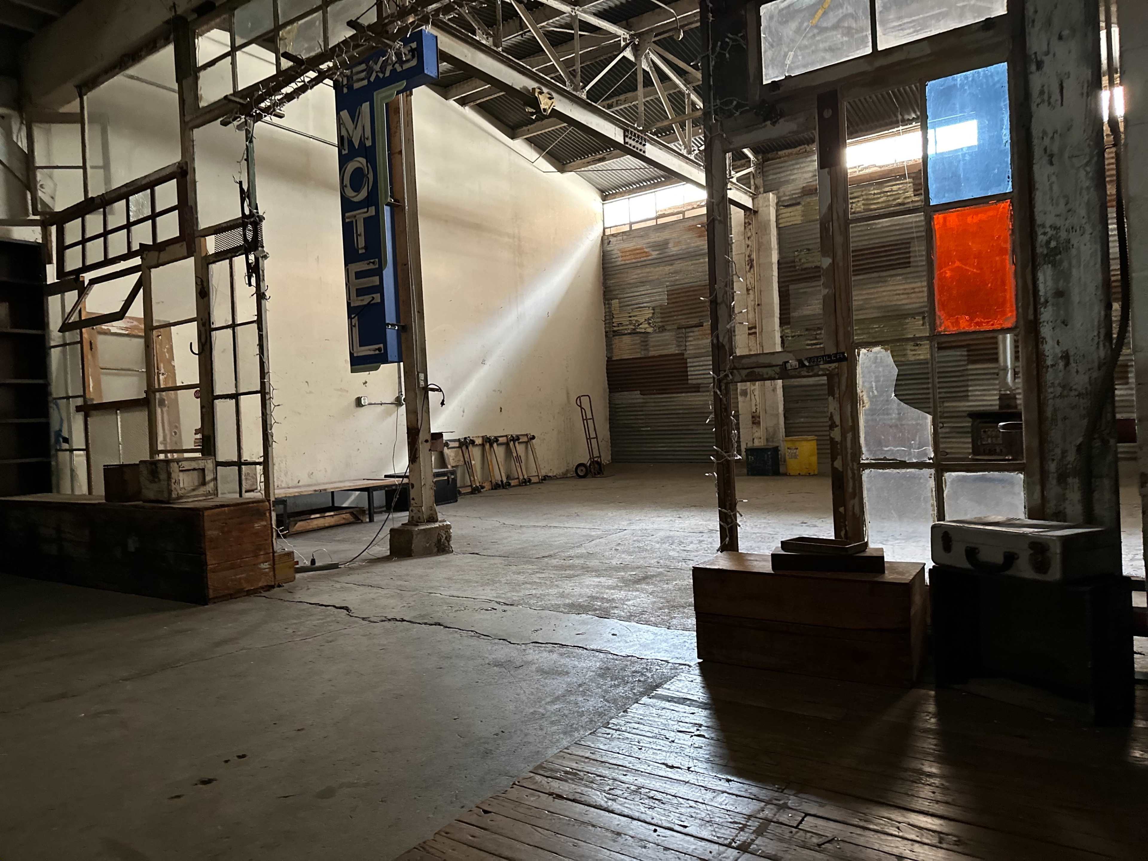 The interior of an empty warehouse features a large "MOTEL" sign and beams of light illuminating scattered wooden structures and a concrete floor.