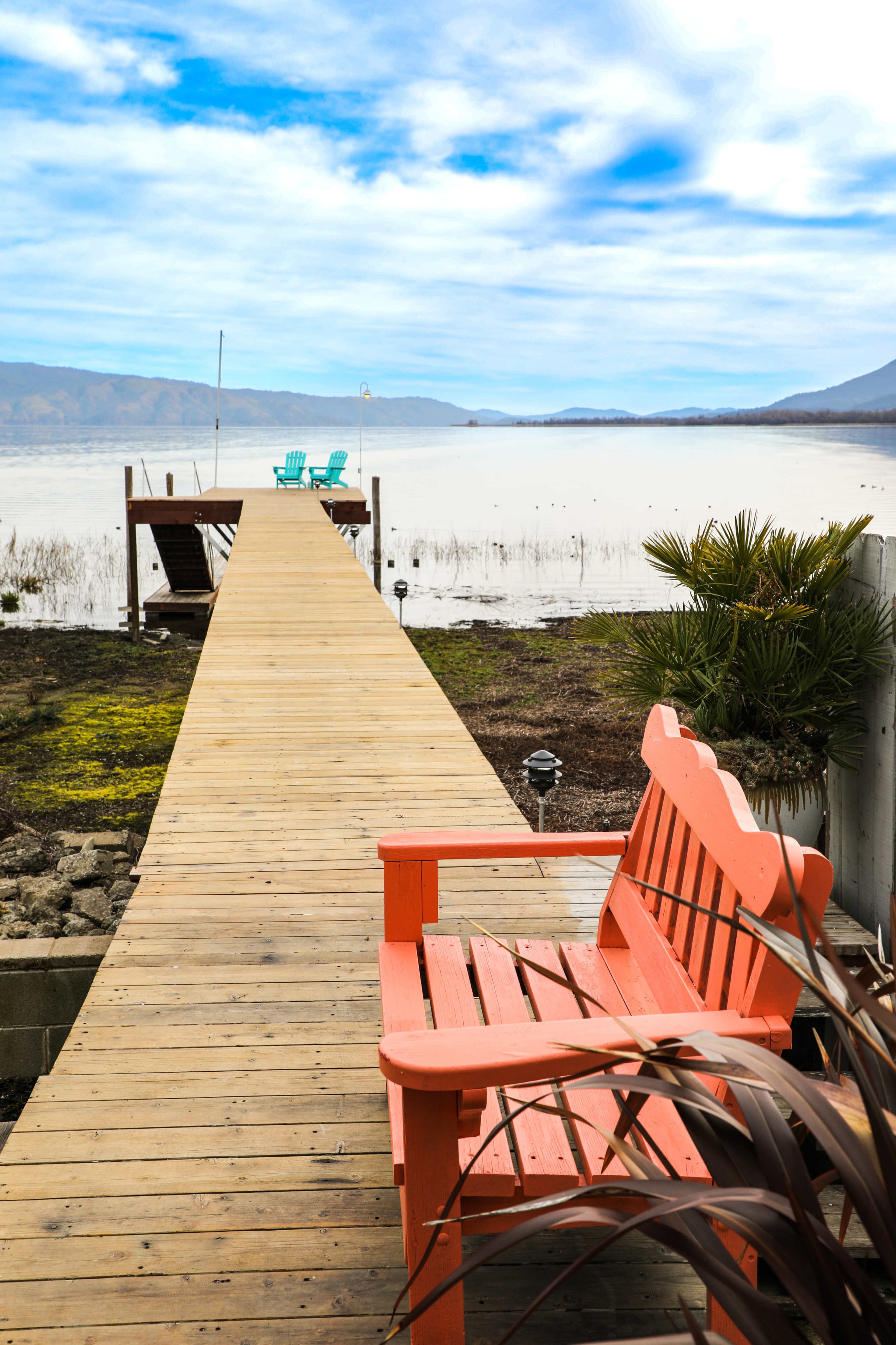 A wooden dock extends into a still lake, with a bright orange bench and blue chairs positioned at the water's edge.