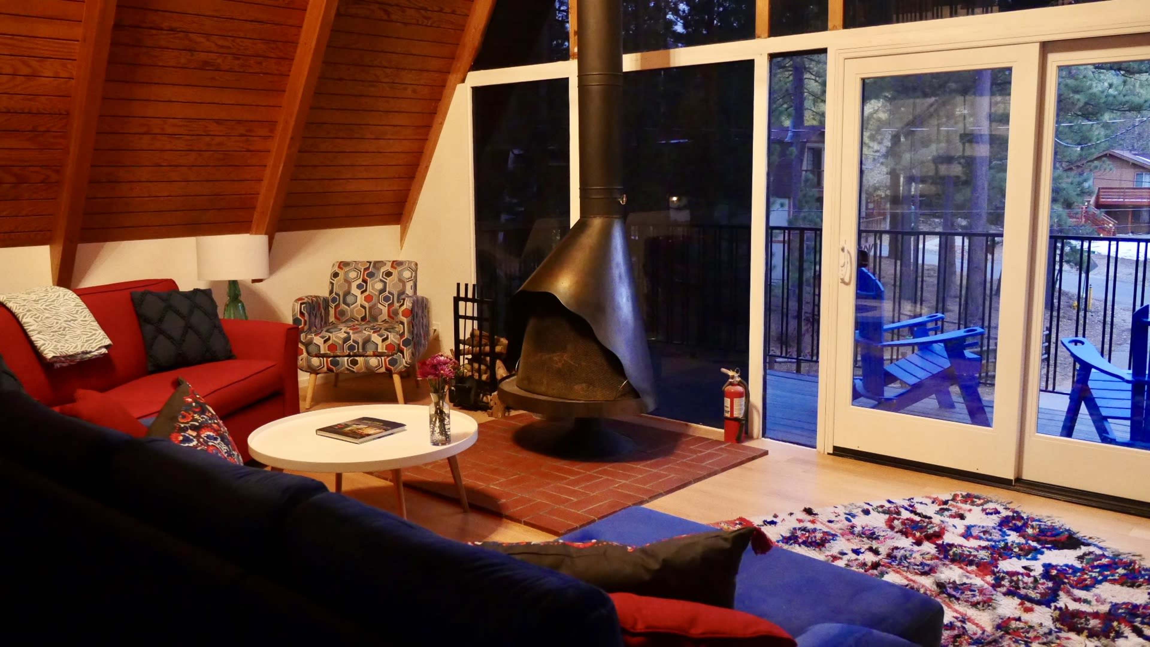 The image shows a cozy living room with a red sofa, a colorful patterned chair, a round coffee table, and a large central fireplace, all set against a backdrop of large glass doors leading to a balcony.