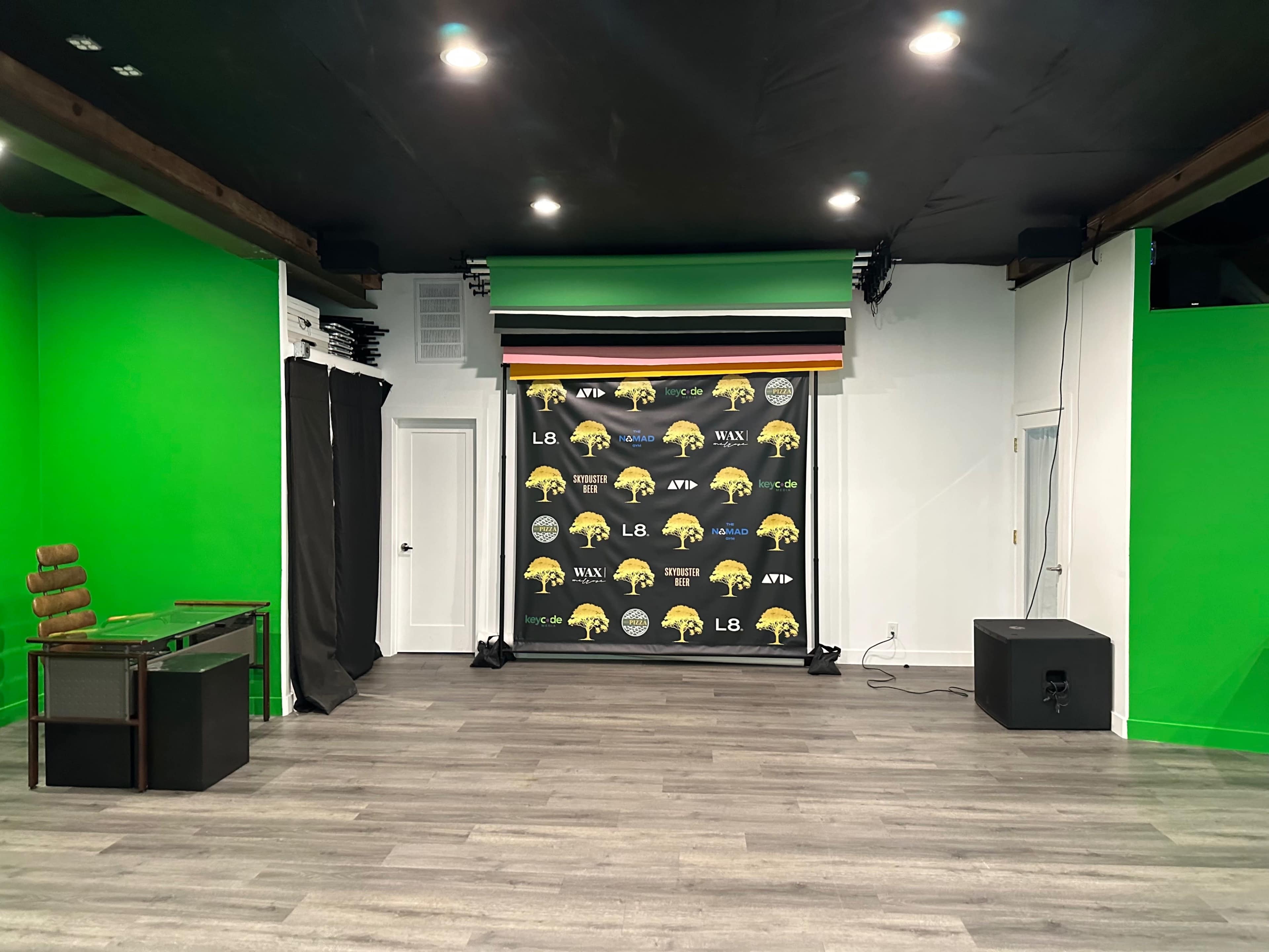 The image shows a studio-like room with a green screen wall, a backdrop featuring promotional graphics, and a table on the left.
