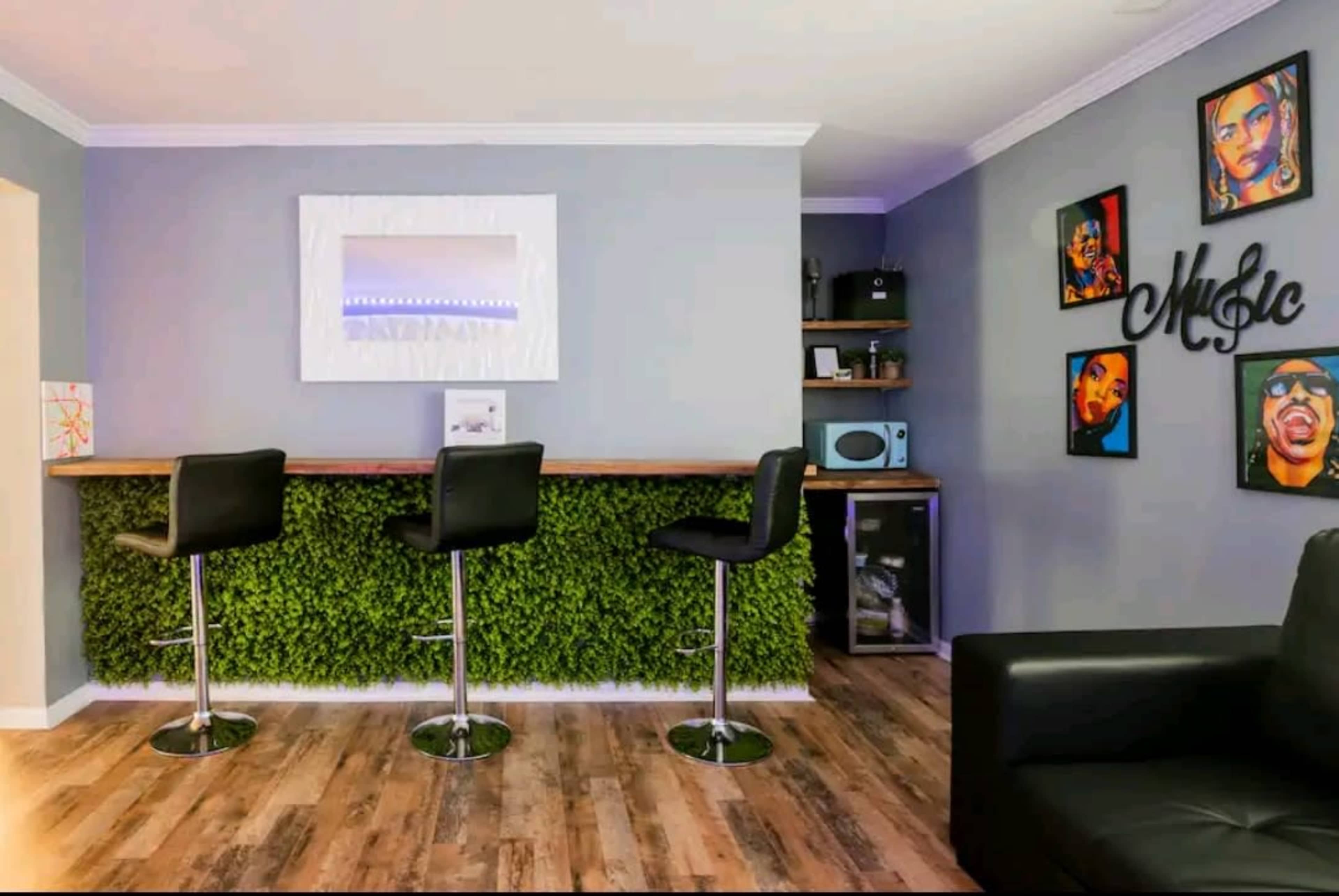 A modern living space with a bar area featuring four black stools, a green wall decoration, and framed artwork of music themes.