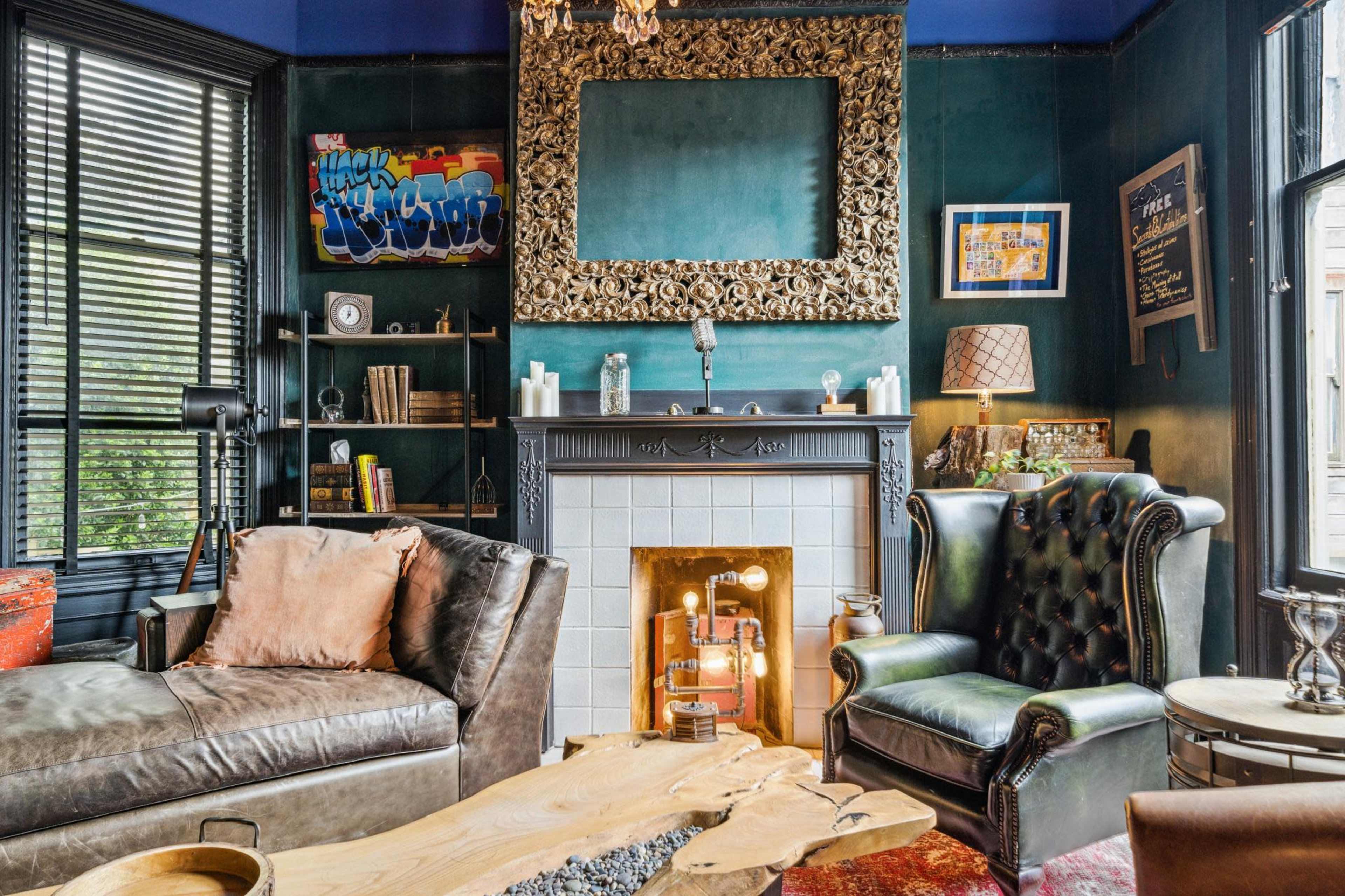 A stylish living room with dark green walls, a decorative fireplace, a leather sofa, and various books and artwork on the shelves.
