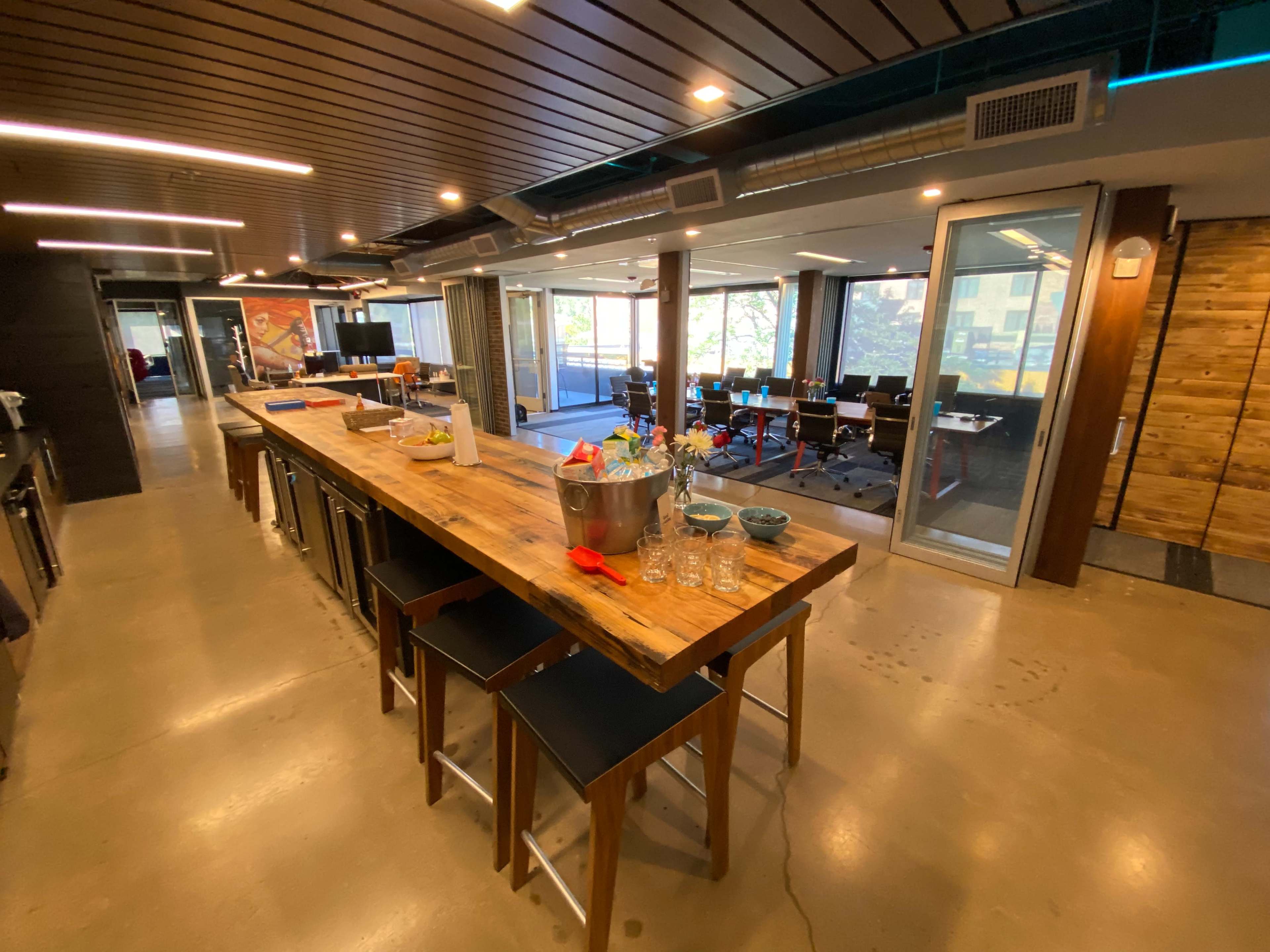 The image shows a modern office kitchen with a large wooden table, stools, and an open area that leads to a conference room filled with chairs and a large window.