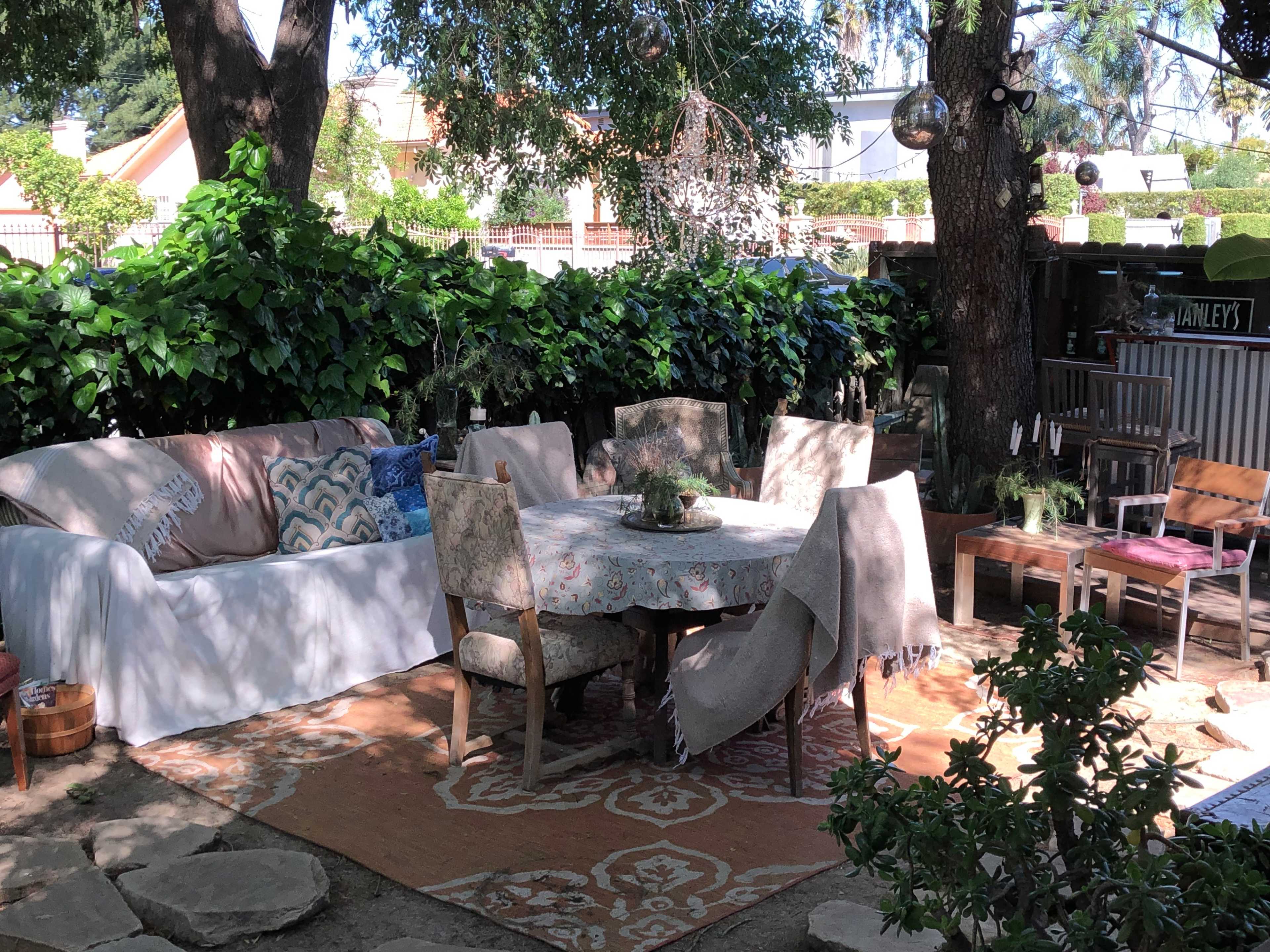 An outdoor seating area with a round table surrounded by chairs, a large white sofa, and greenery in the background.