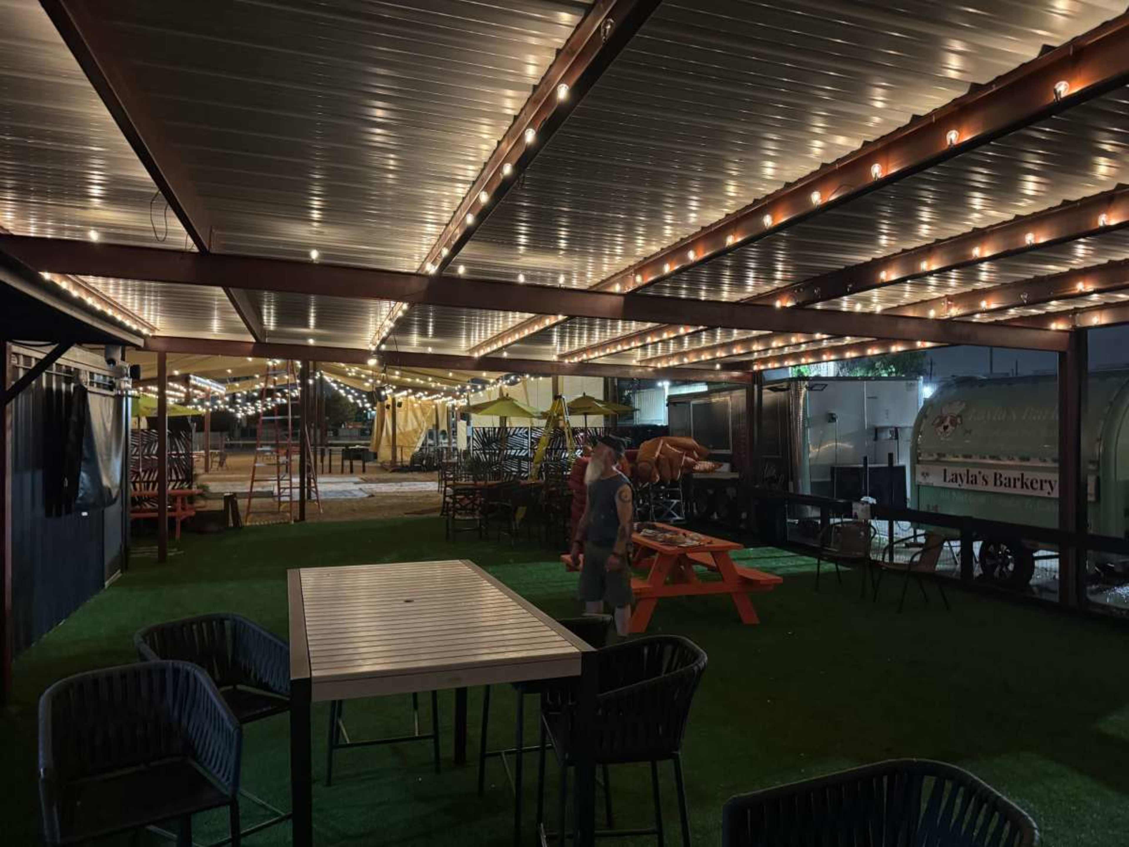 The image shows a dimly lit outdoor seating area with string lights overhead, featuring tables and chairs arranged on a grassy surface.