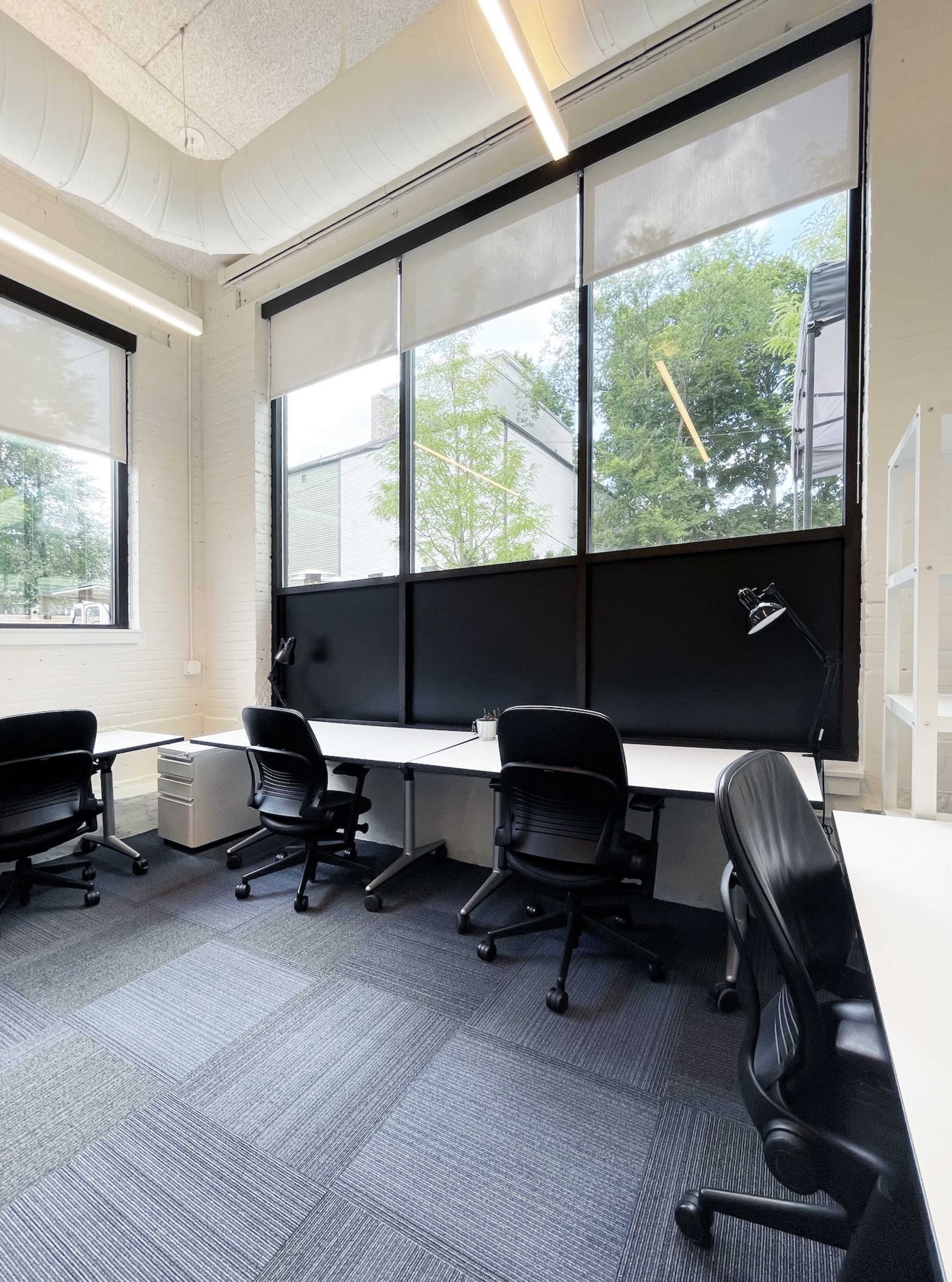 A modern office space with several black chairs positioned at white desks beneath large windows.