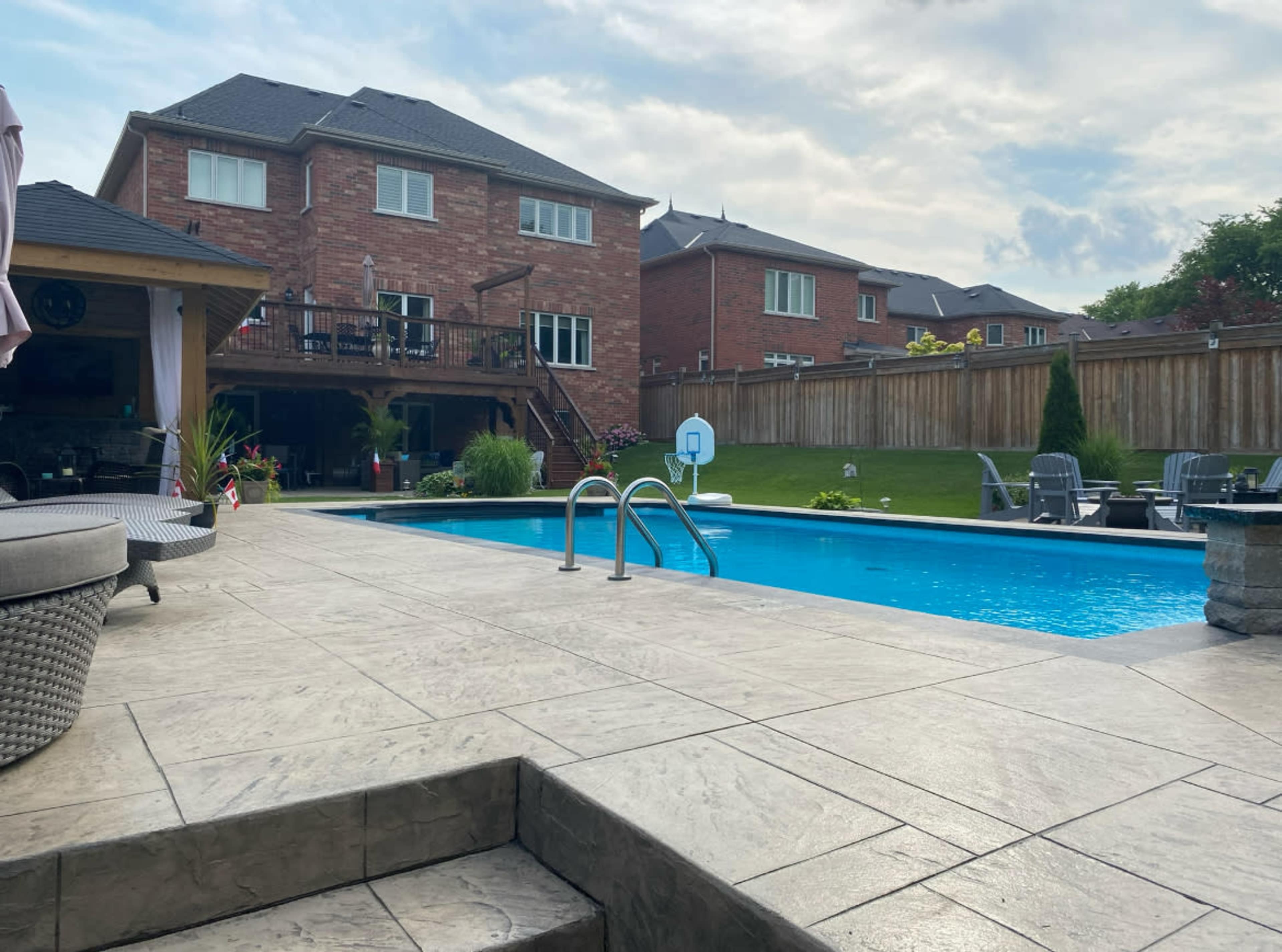 A backyard features a swimming pool surrounded by a stone patio and lounge chairs, with a two-story brick house in the background.