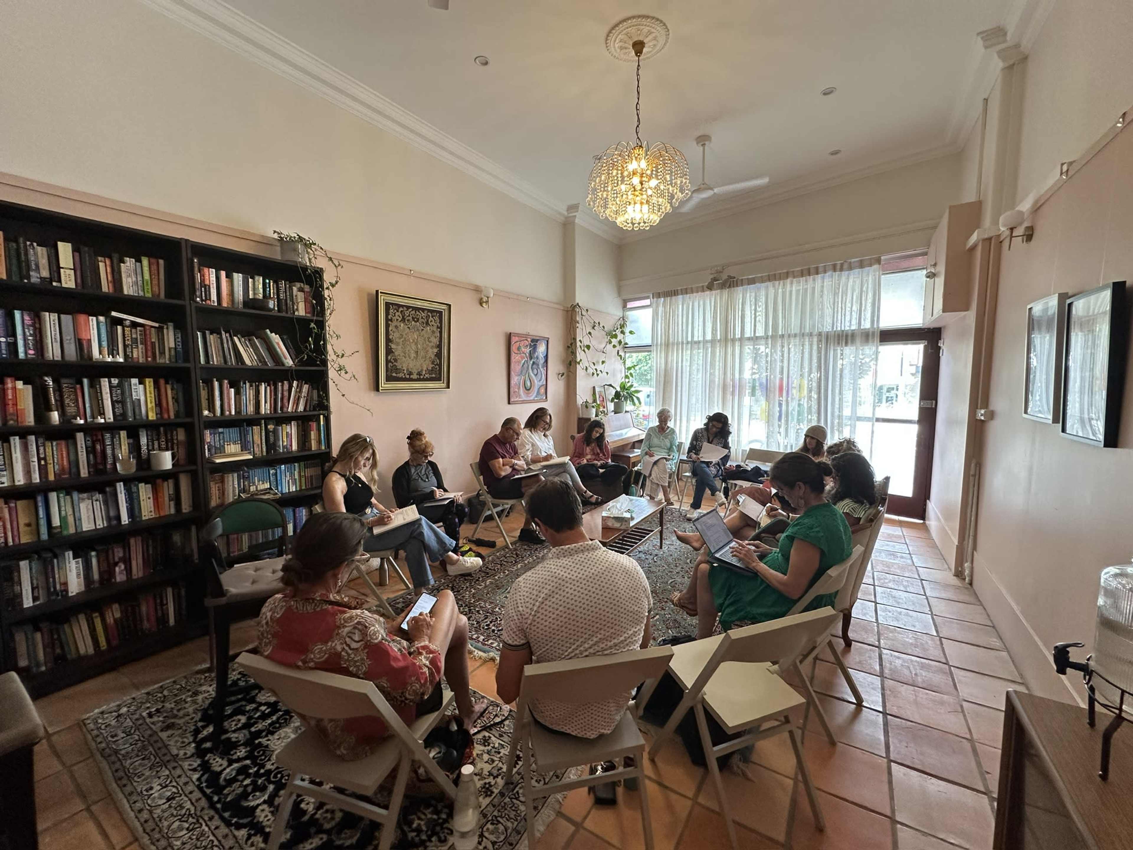 A group of people sits in a circle on folding chairs in a well-lit room filled with bookshelves and decorative plants.