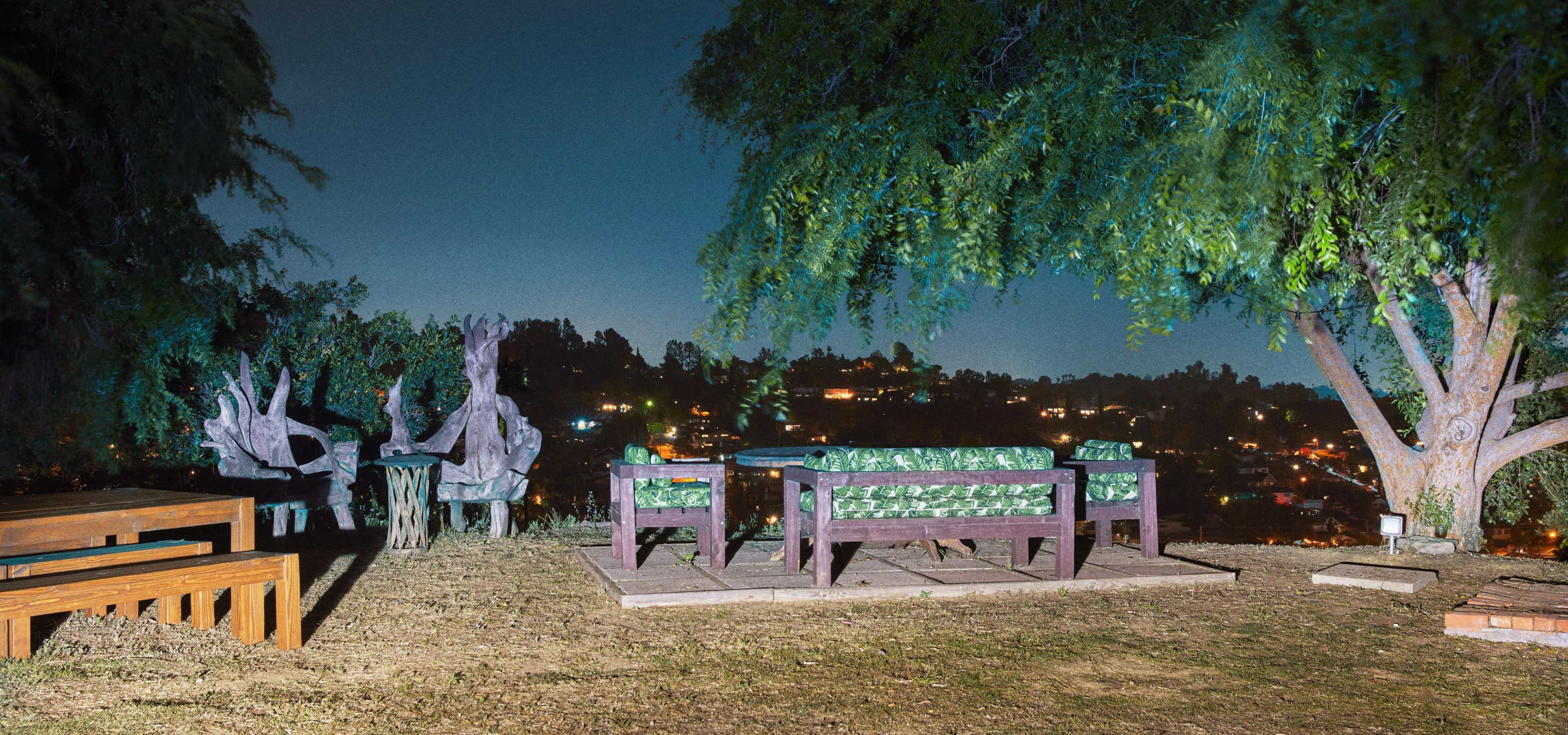 The scene shows outdoor seating arrangements on a grassy area with a view of a city skyline at night, illuminated by various lights.