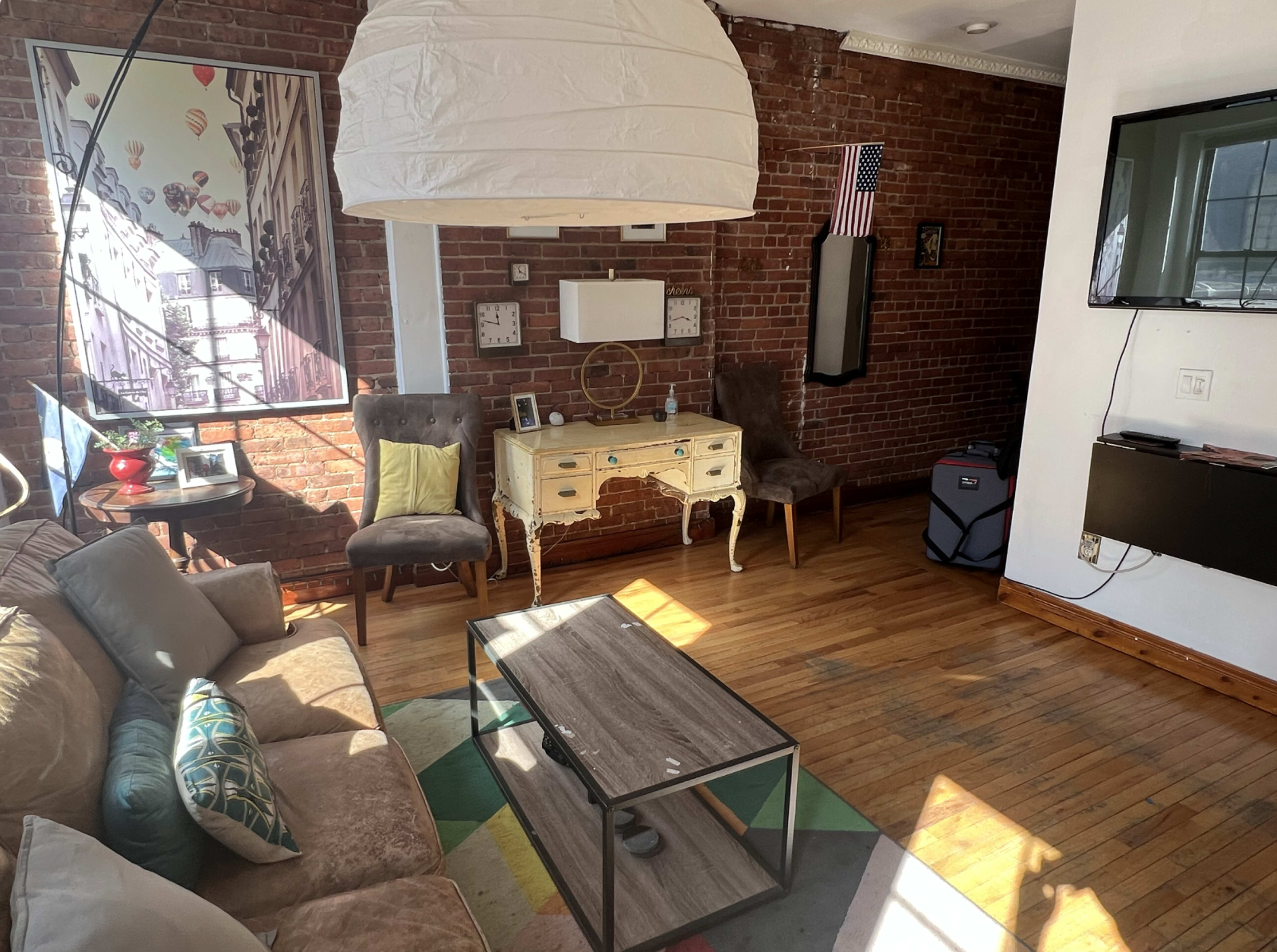 A living room with exposed brick walls, a couch, two chairs, a coffee table, and a decorative white light fixture hanging from the ceiling.