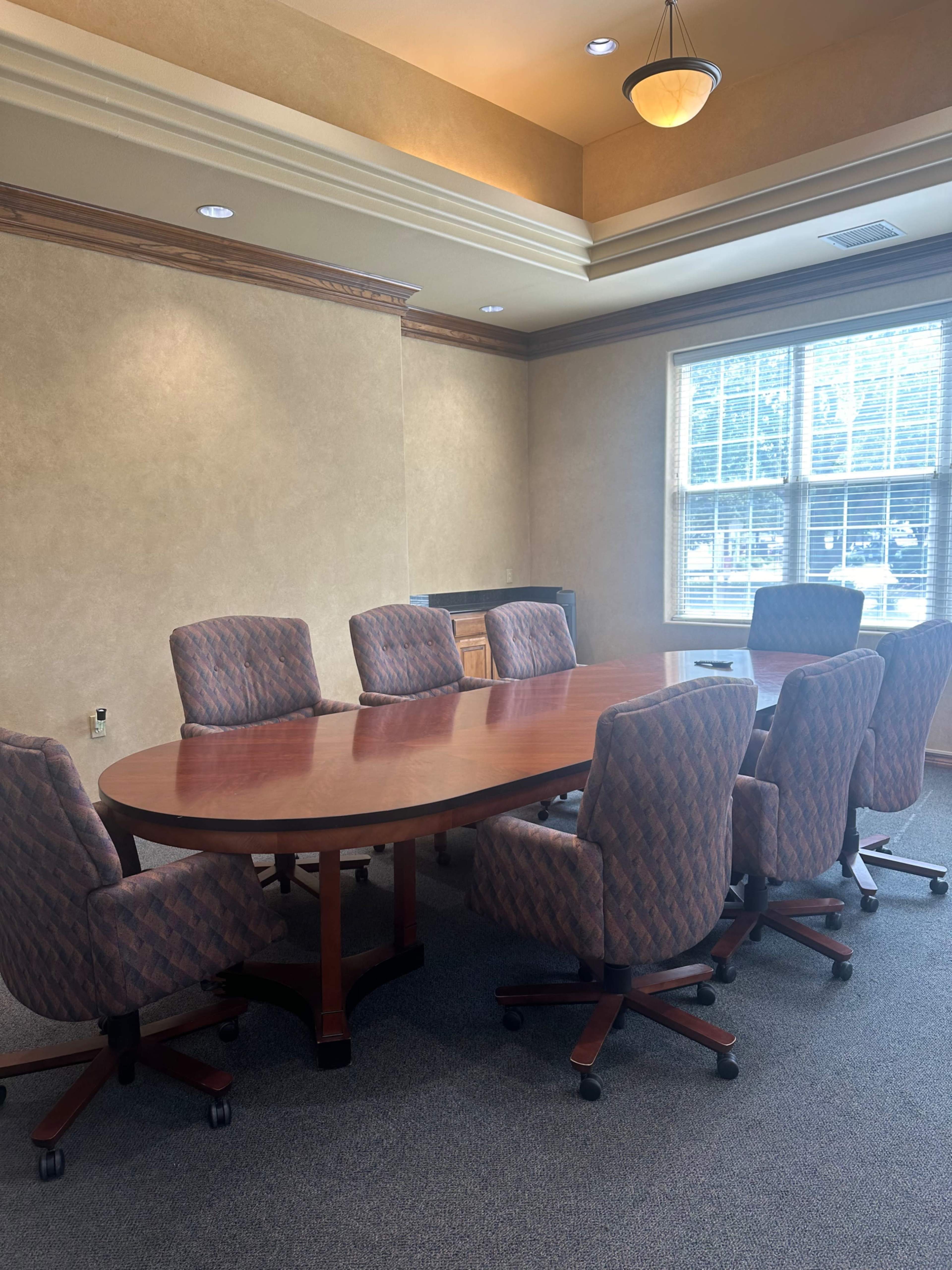 The image shows a conference room with a large oval wooden table surrounded by eight upholstered chairs.