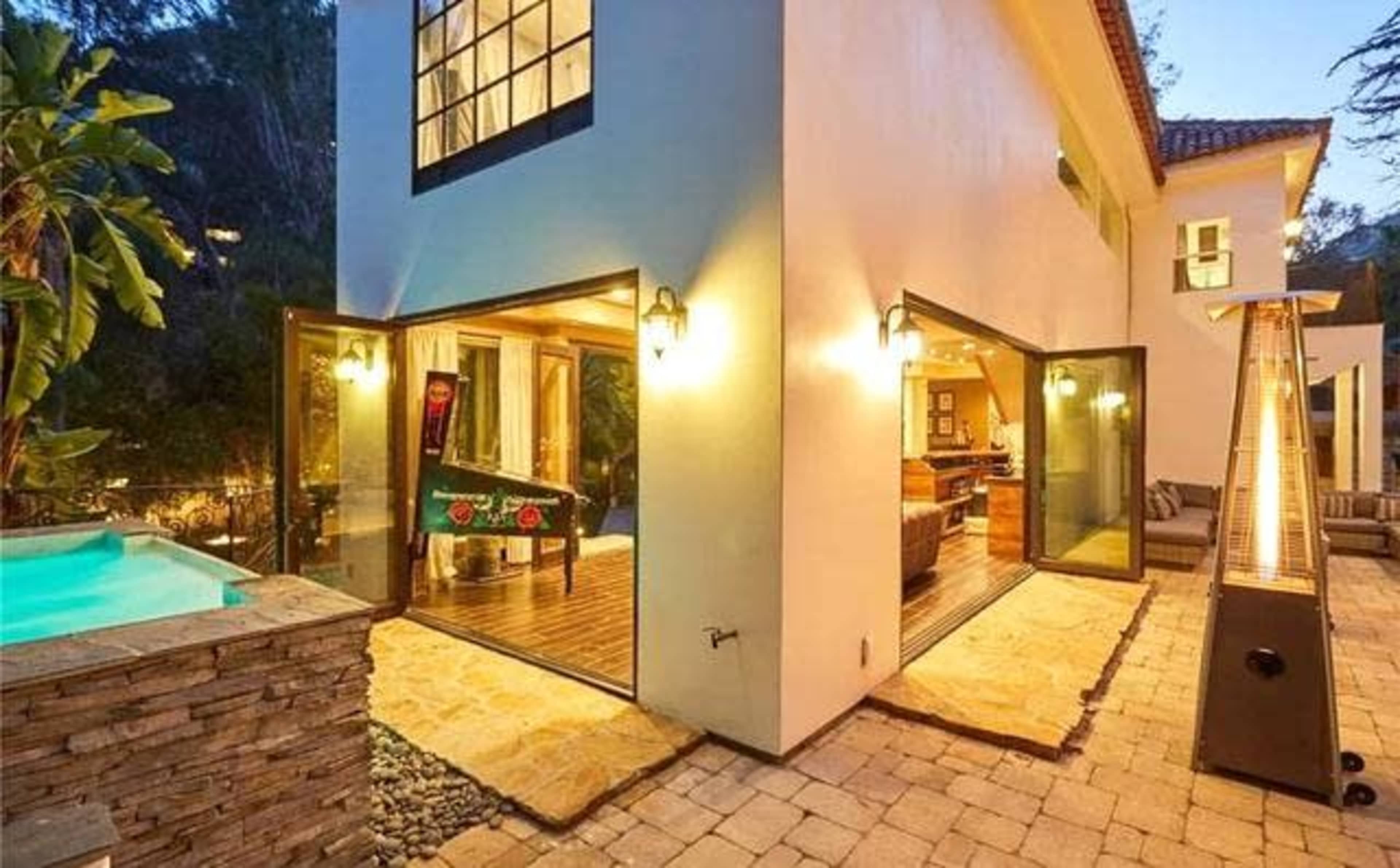 An outdoor area features a pool, a game table, and a patio with a stone wall and heating lamps, adjoining a modern home.