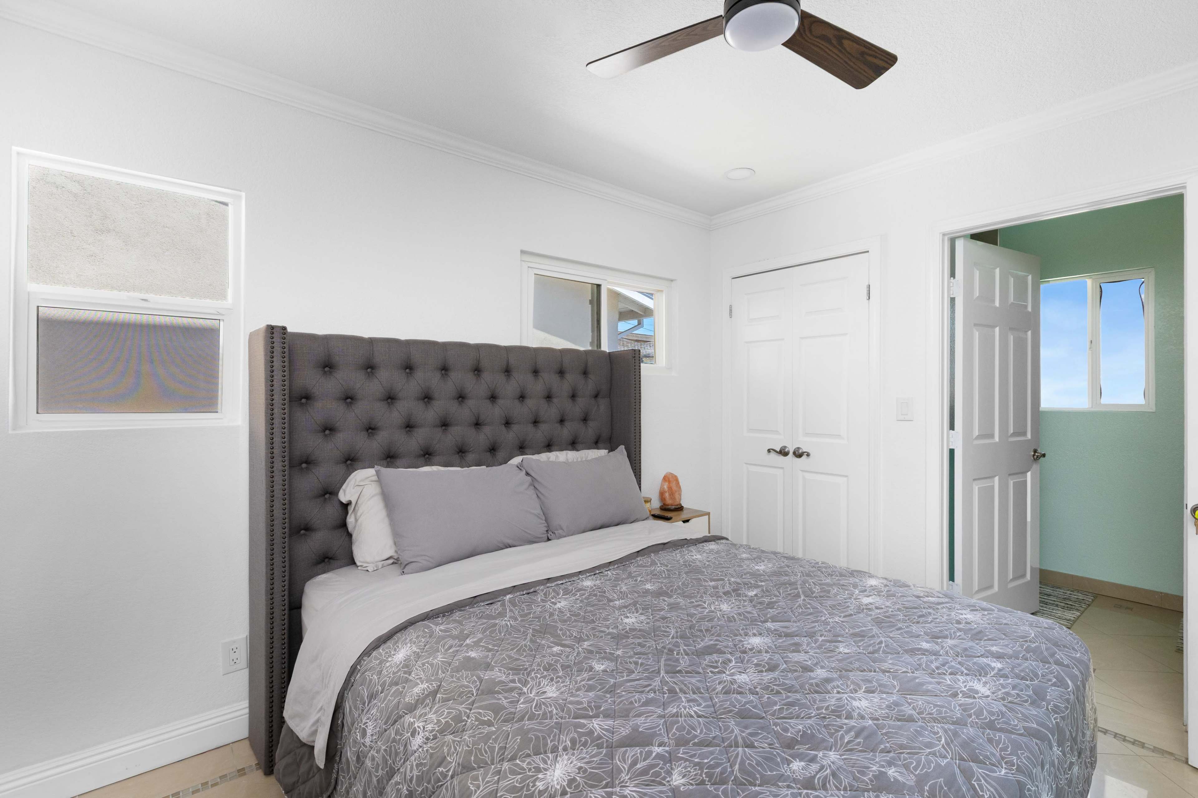 The image shows a modern bedroom with a tufted gray bed frame, a light gray blanket, and two windows allowing natural light to enter the room.