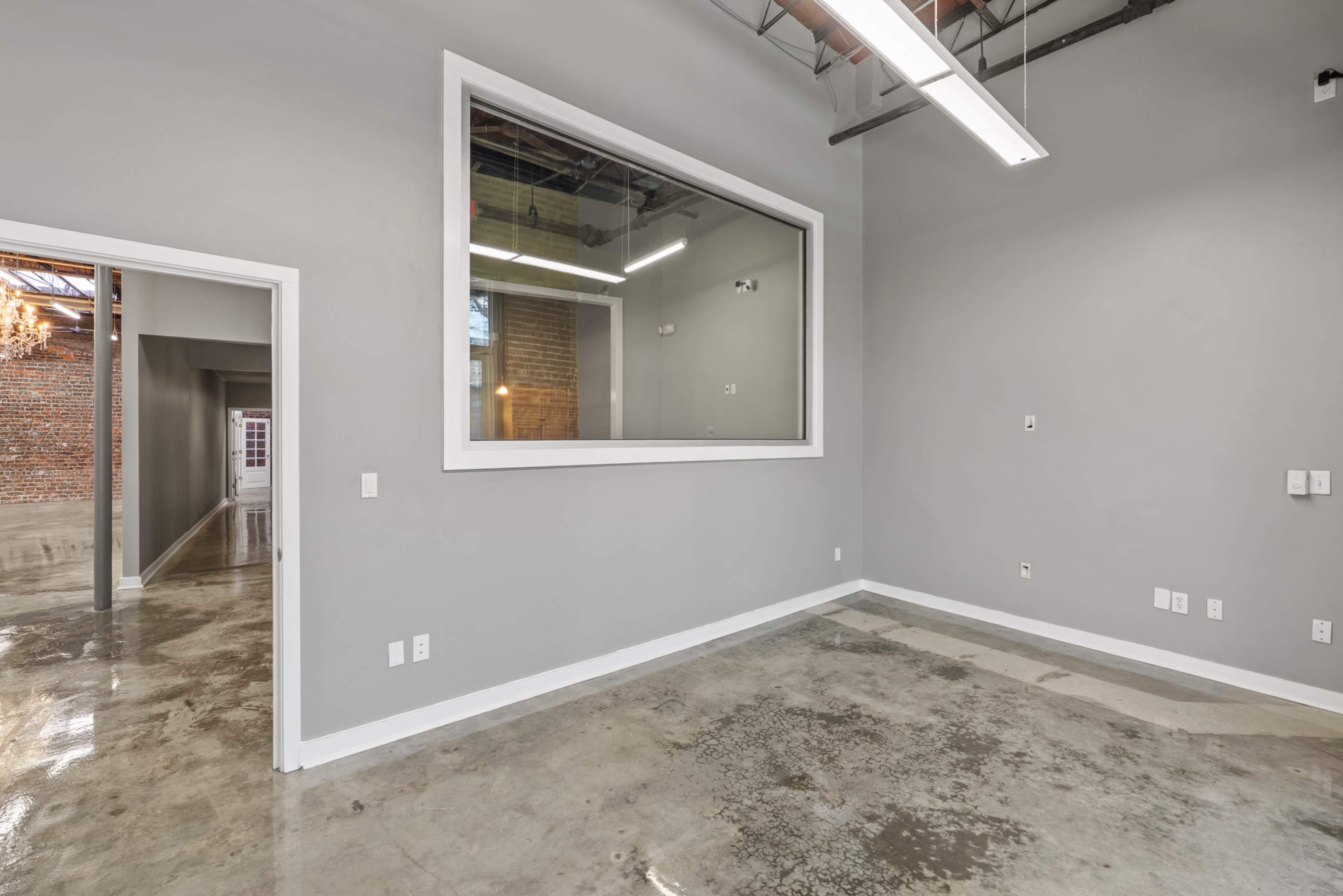 The image shows a spacious, empty room with gray walls, polished concrete flooring, and a large window that overlooks another area.