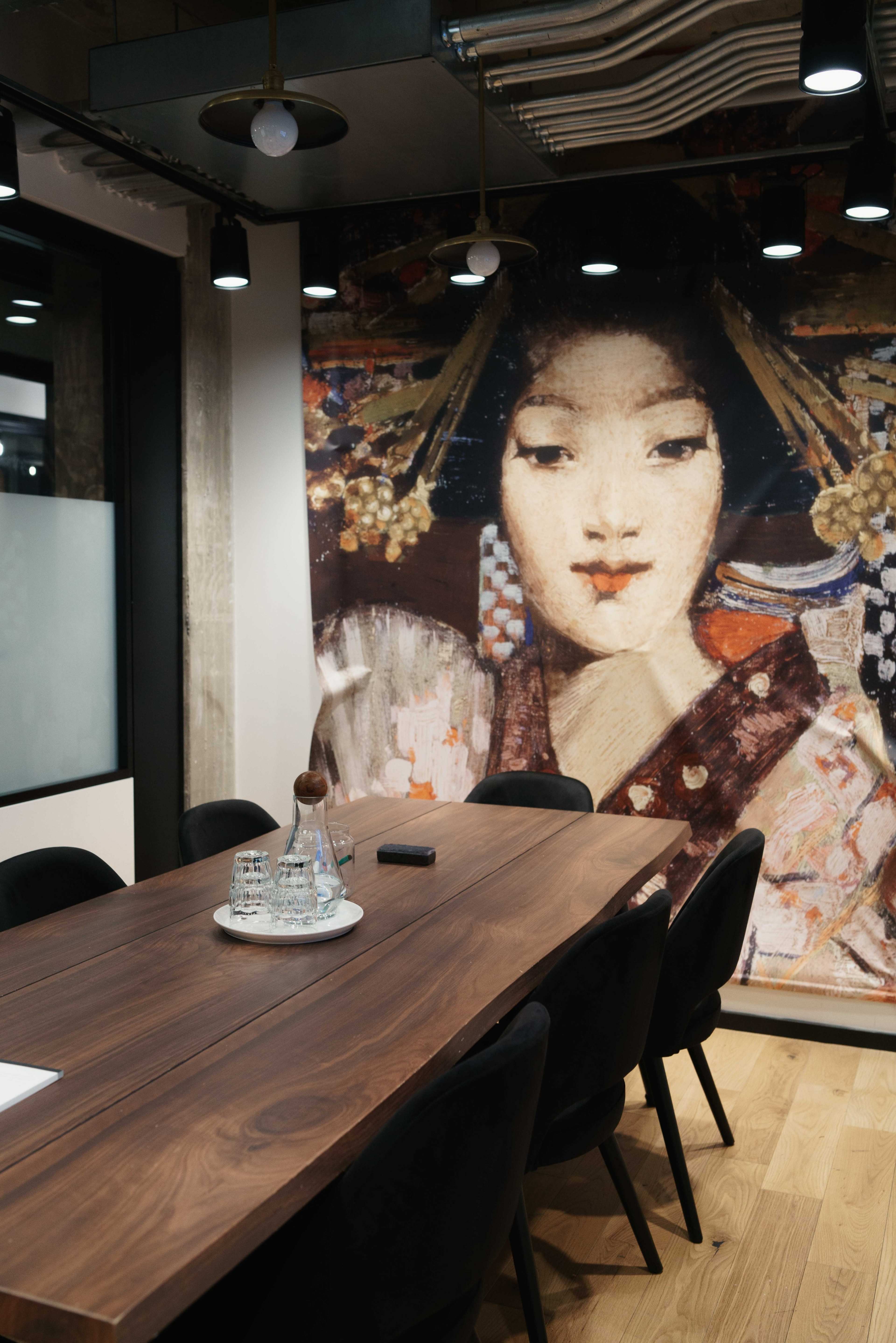 A modern conference room features a long wooden table surrounded by black chairs, with a traditional Japanese art piece displayed on the wall.