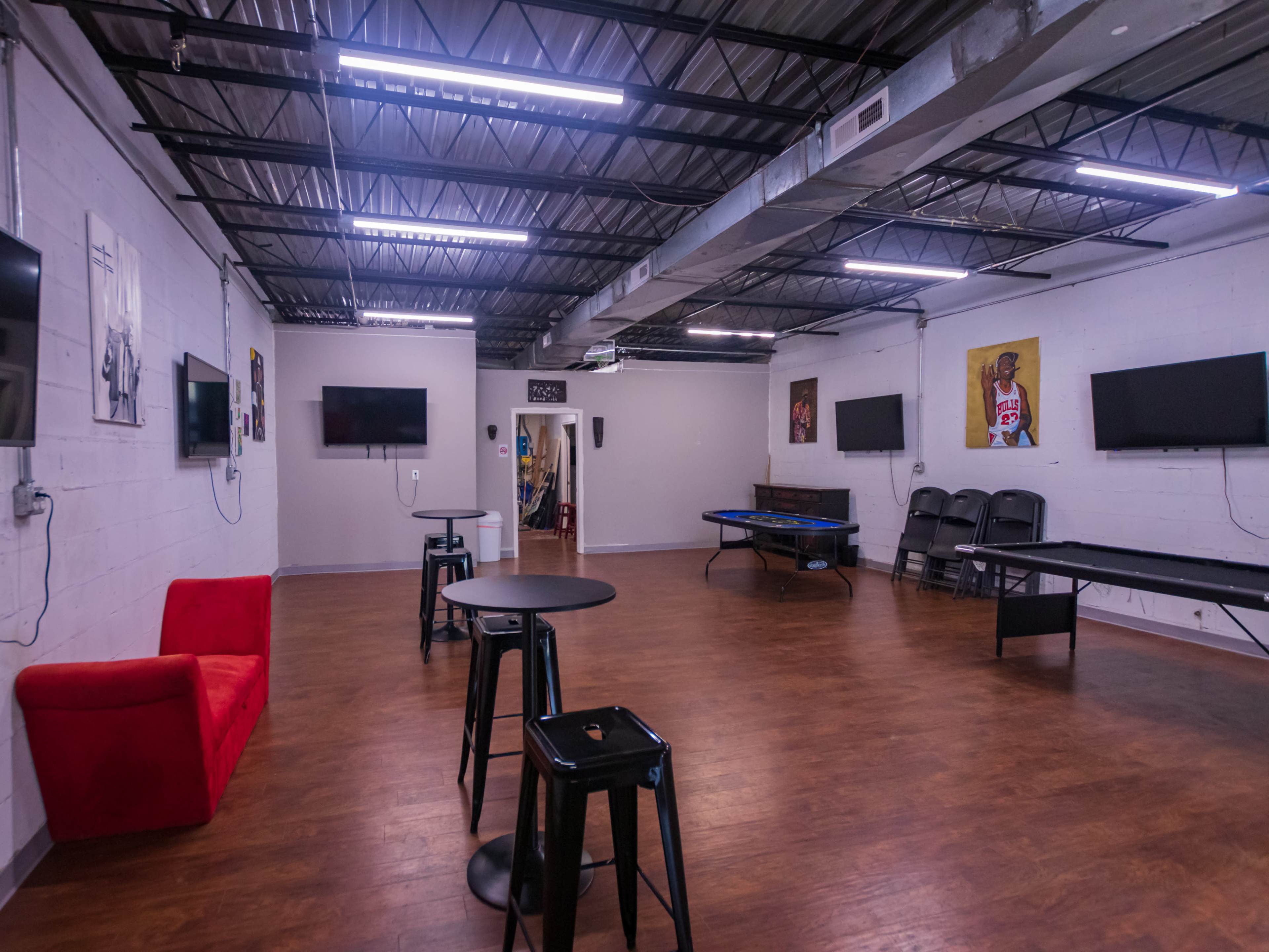 A spacious room with wood flooring, featuring a red couch, several tables and chairs, wall-mounted televisions, and artwork on the walls.