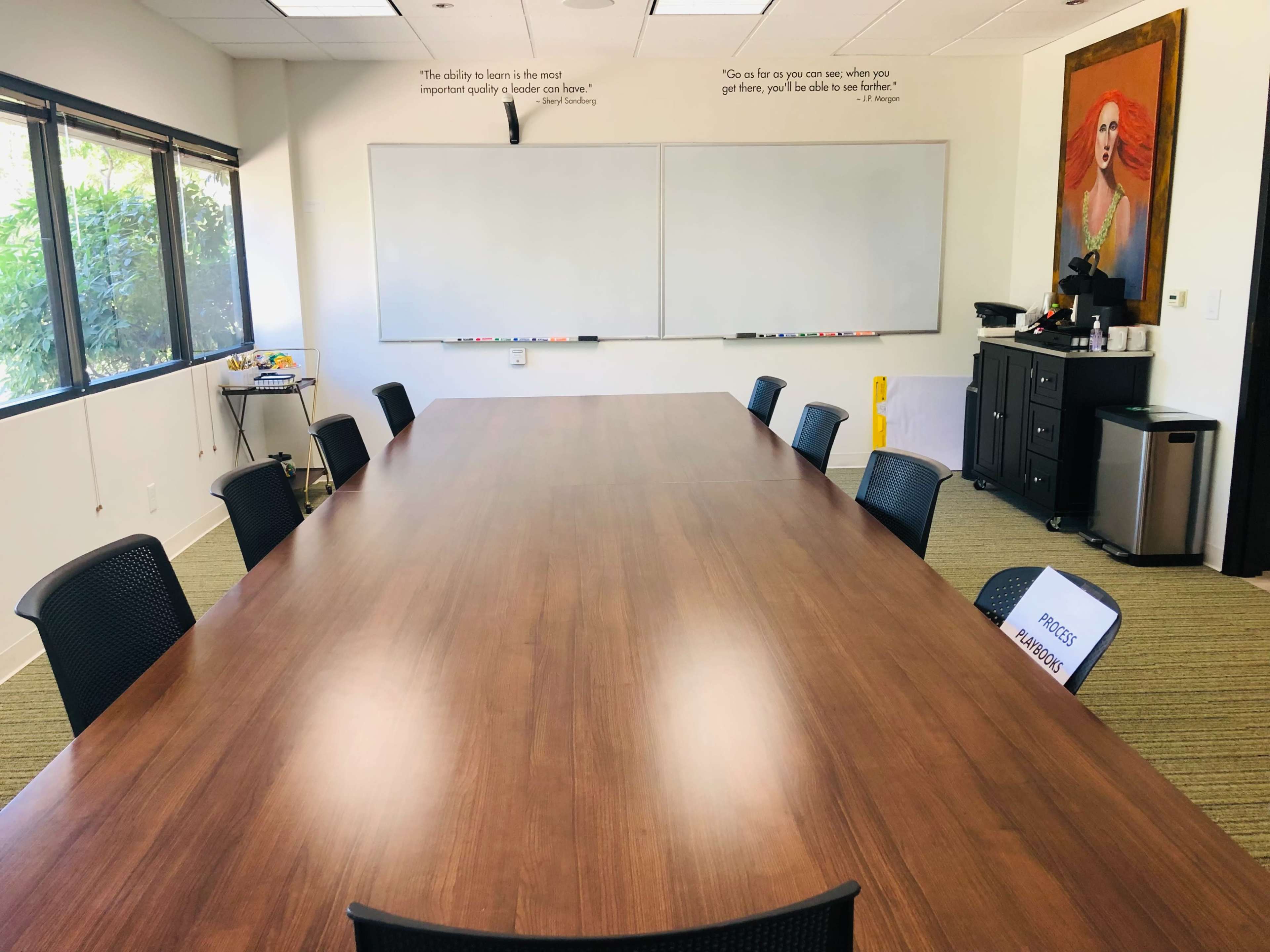 An empty conference room features a long wooden table surrounded by black chairs, with whiteboards on the walls and a coffee station in the corner.