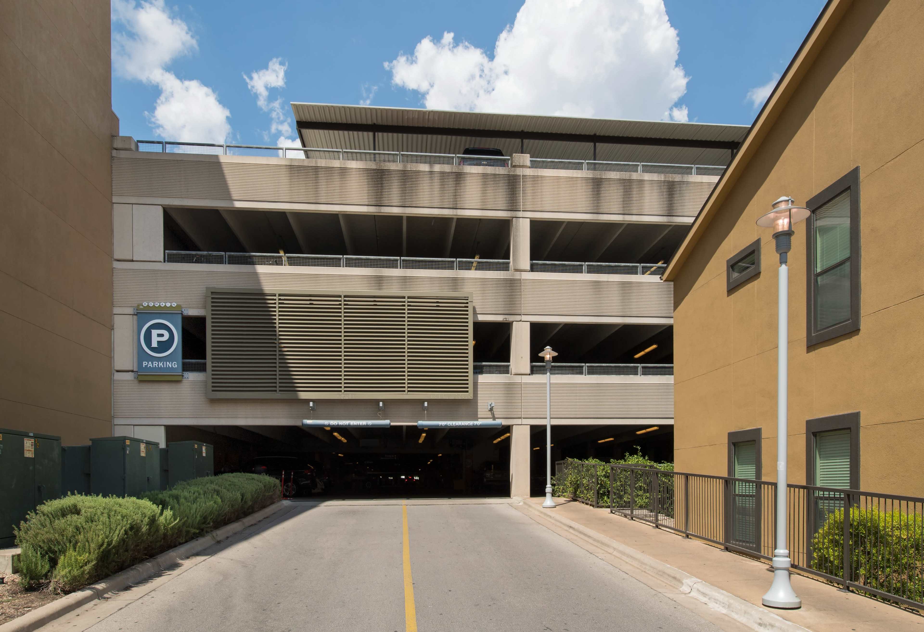 A multi-level parking garage is situated at the end of a driveway, flanked by greenery and a building.