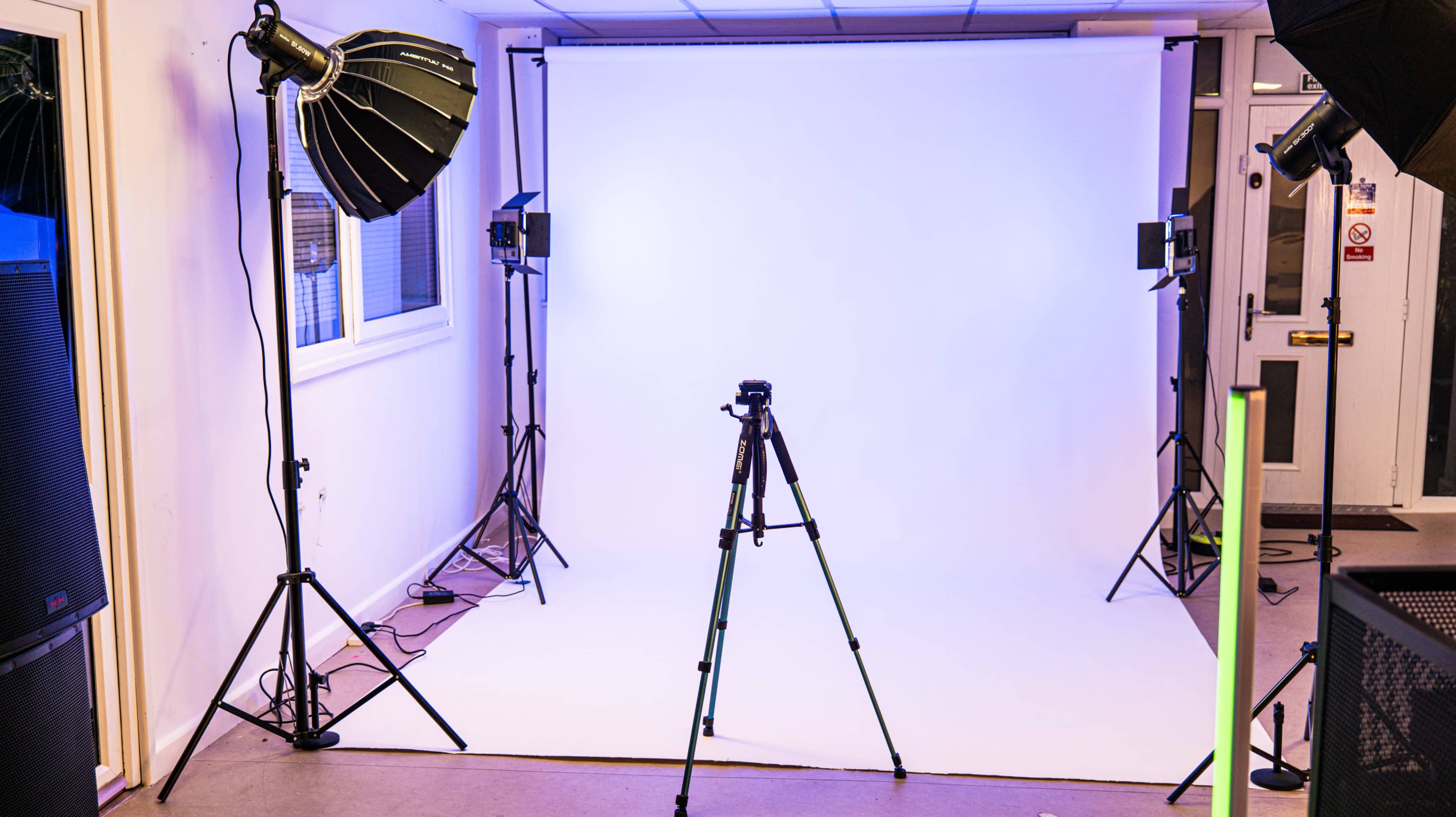 The image shows a photography studio setup with a white backdrop, a camera tripod in the foreground, and several lighting equipment positioned around the space.