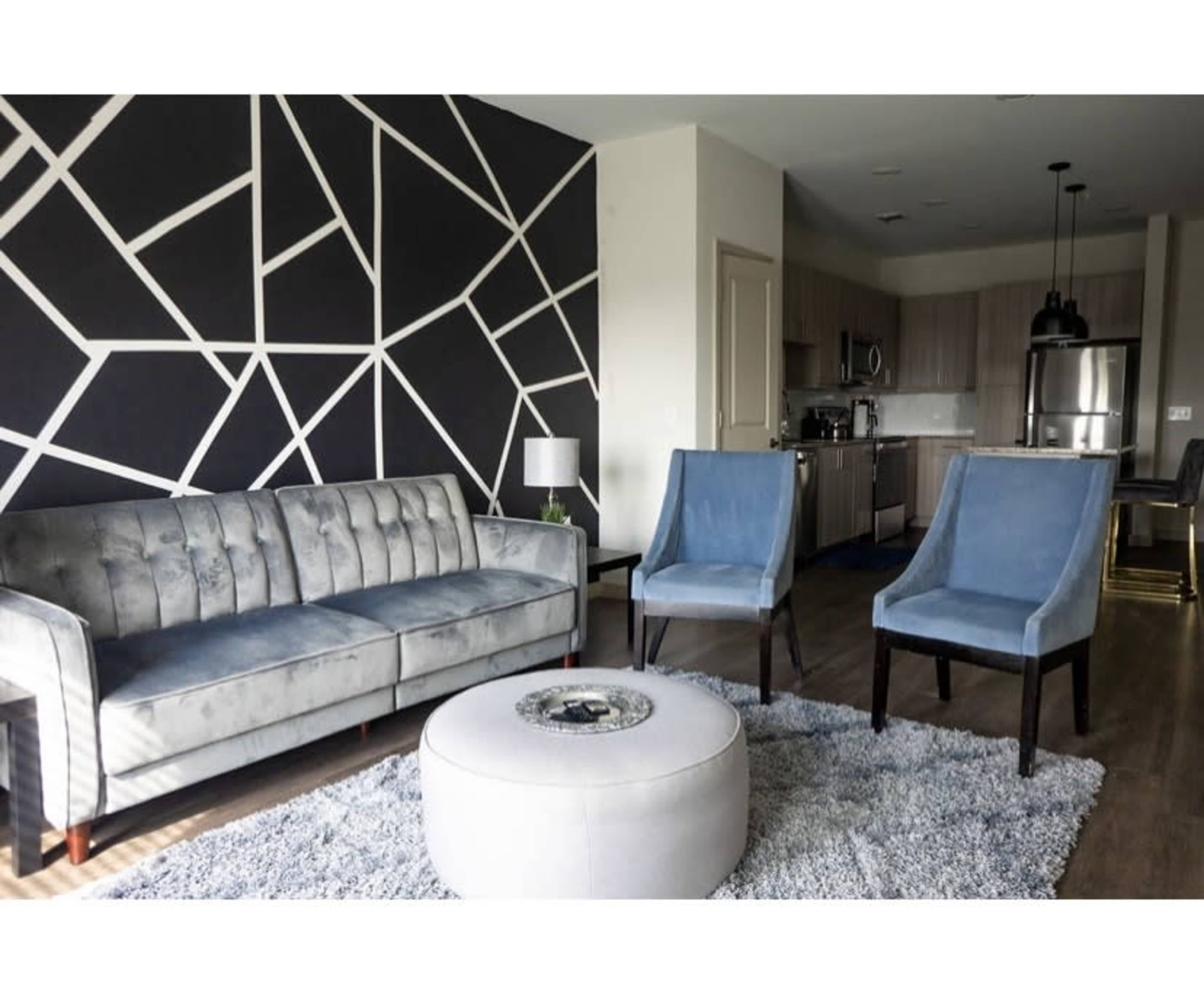 A modern living room featuring a gray sofa, two blue velvet chairs, a round coffee table, and a geometric wall design.