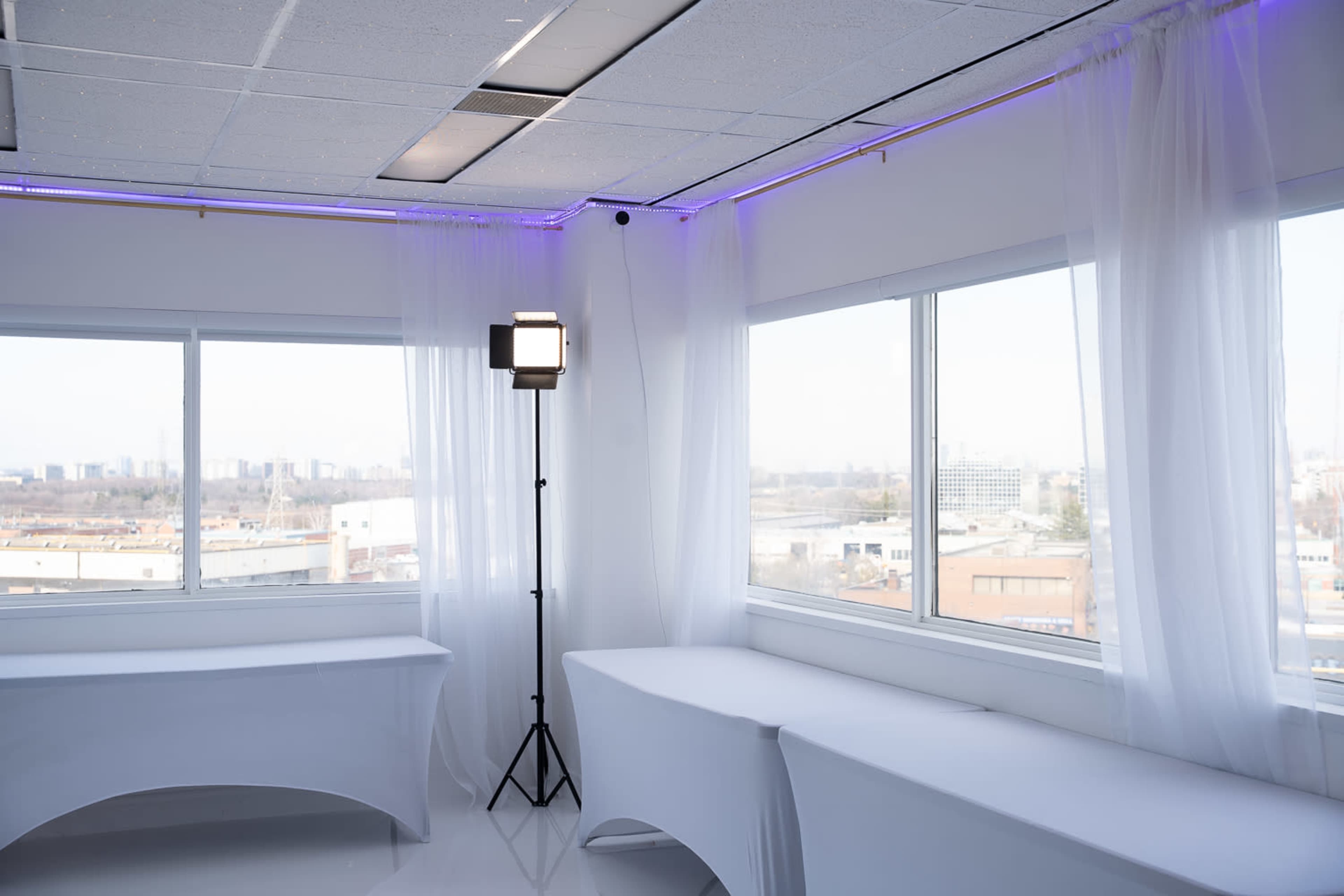 The image shows a bright, modern room with large windows, white drapes, and a lighting setup on a stand.