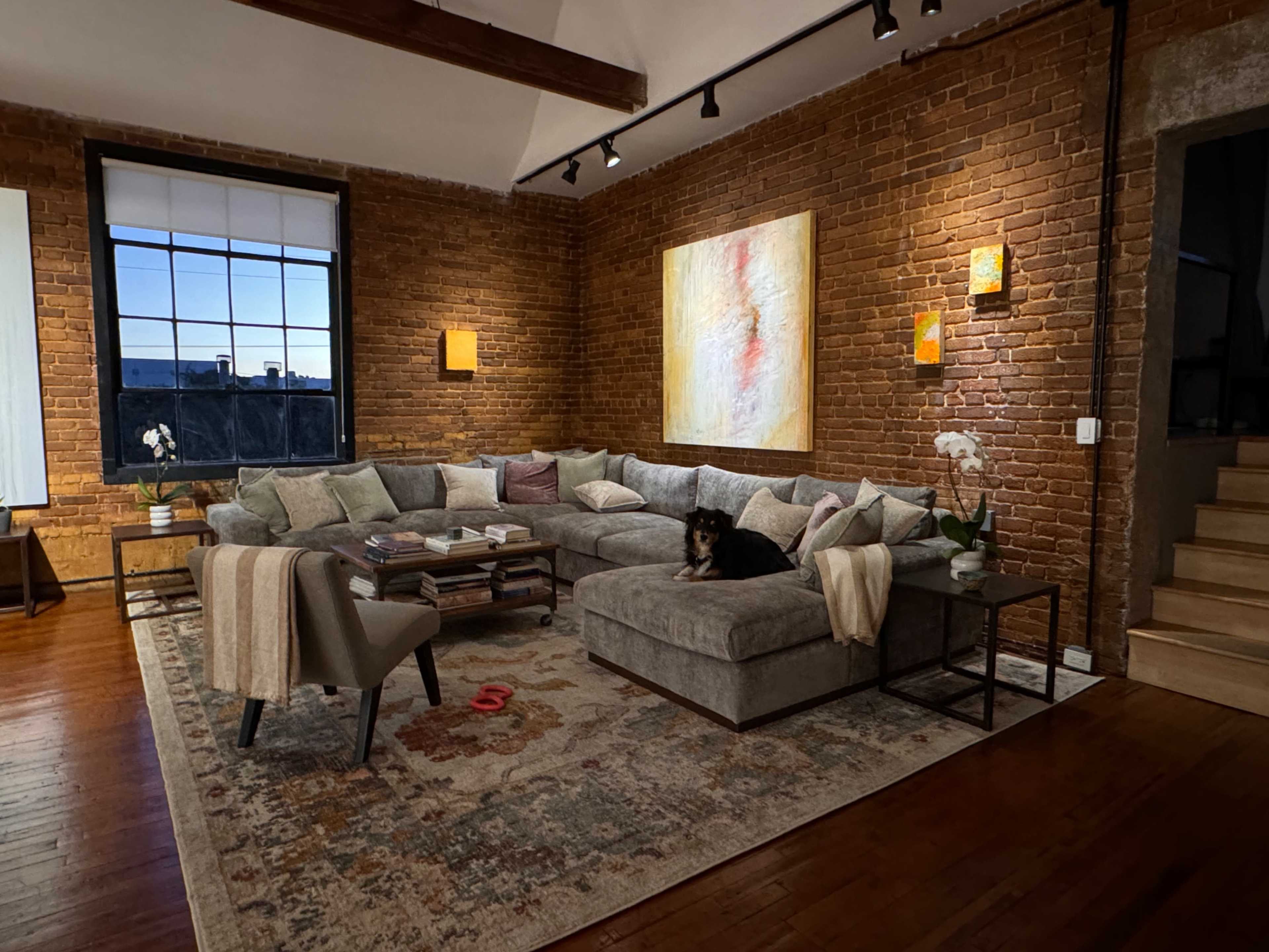 A spacious living room features a large sectional sofa, a coffee table with various items, two wall-mounted lights, and an abstract painting on a brick wall.