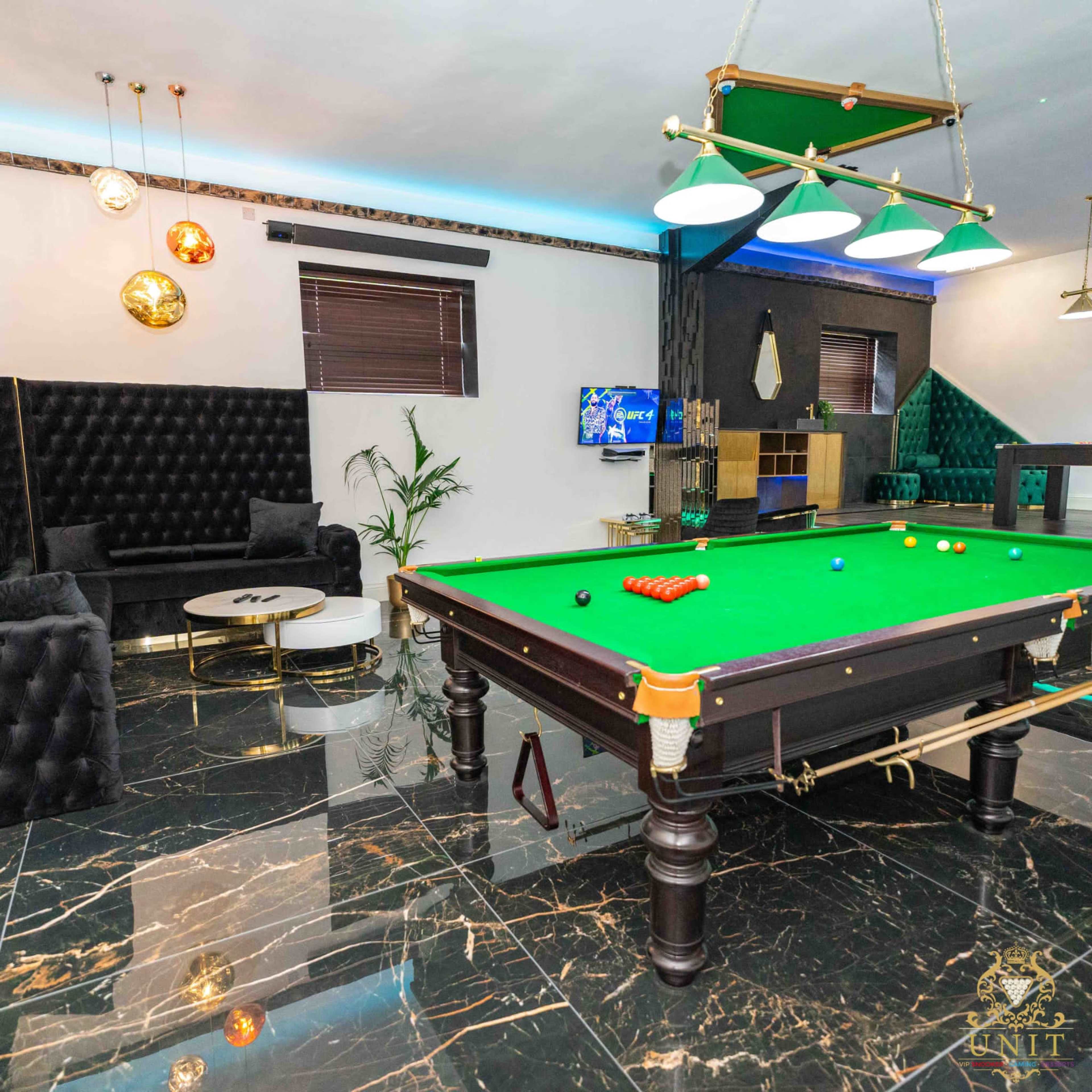 The image shows a billiards room featuring a pool table with a green felt surface, surrounded by modern seating and decorative lighting.