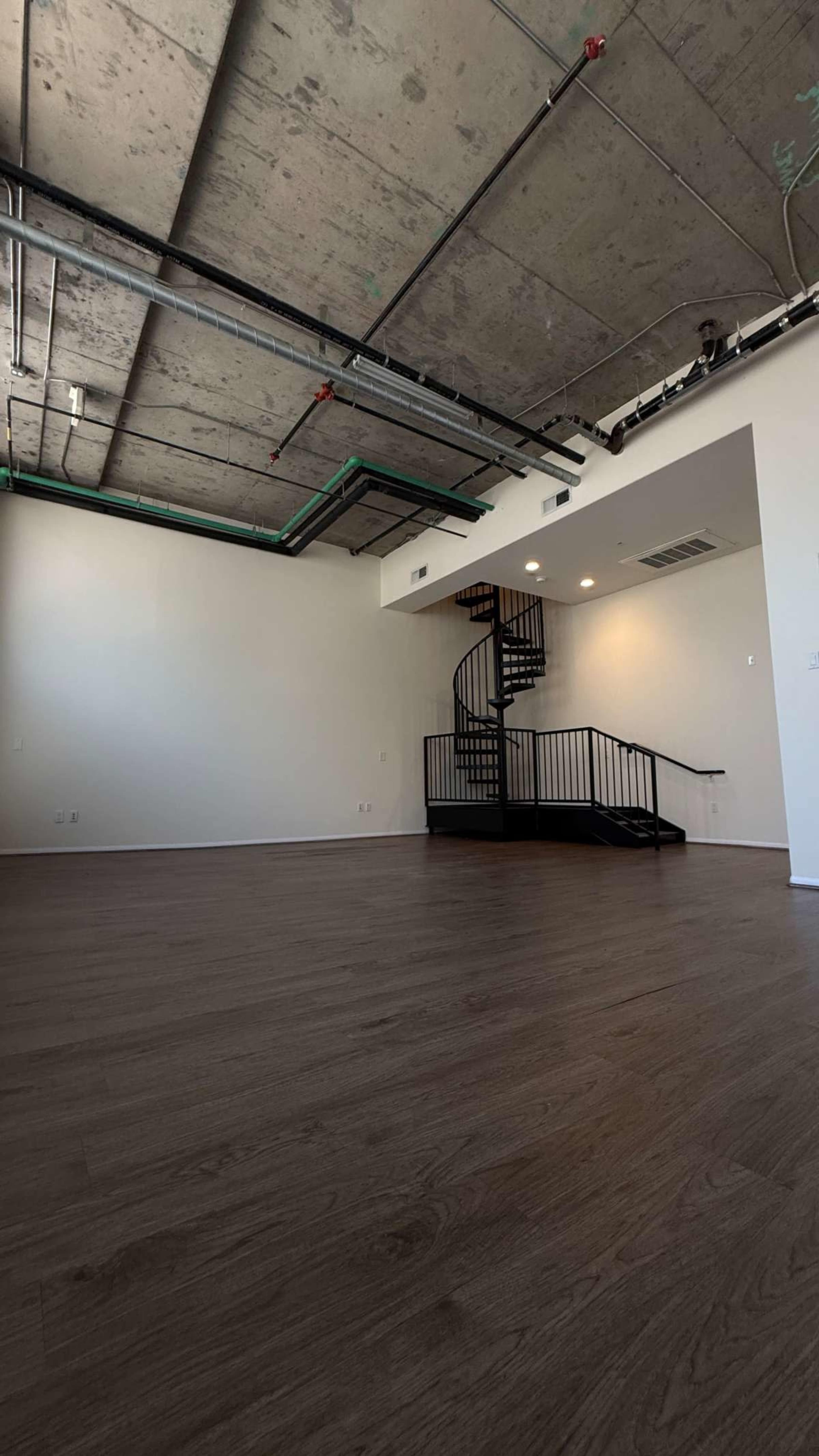 The image shows an empty room with wooden flooring and a spiral staircase along the far wall under a concrete ceiling.