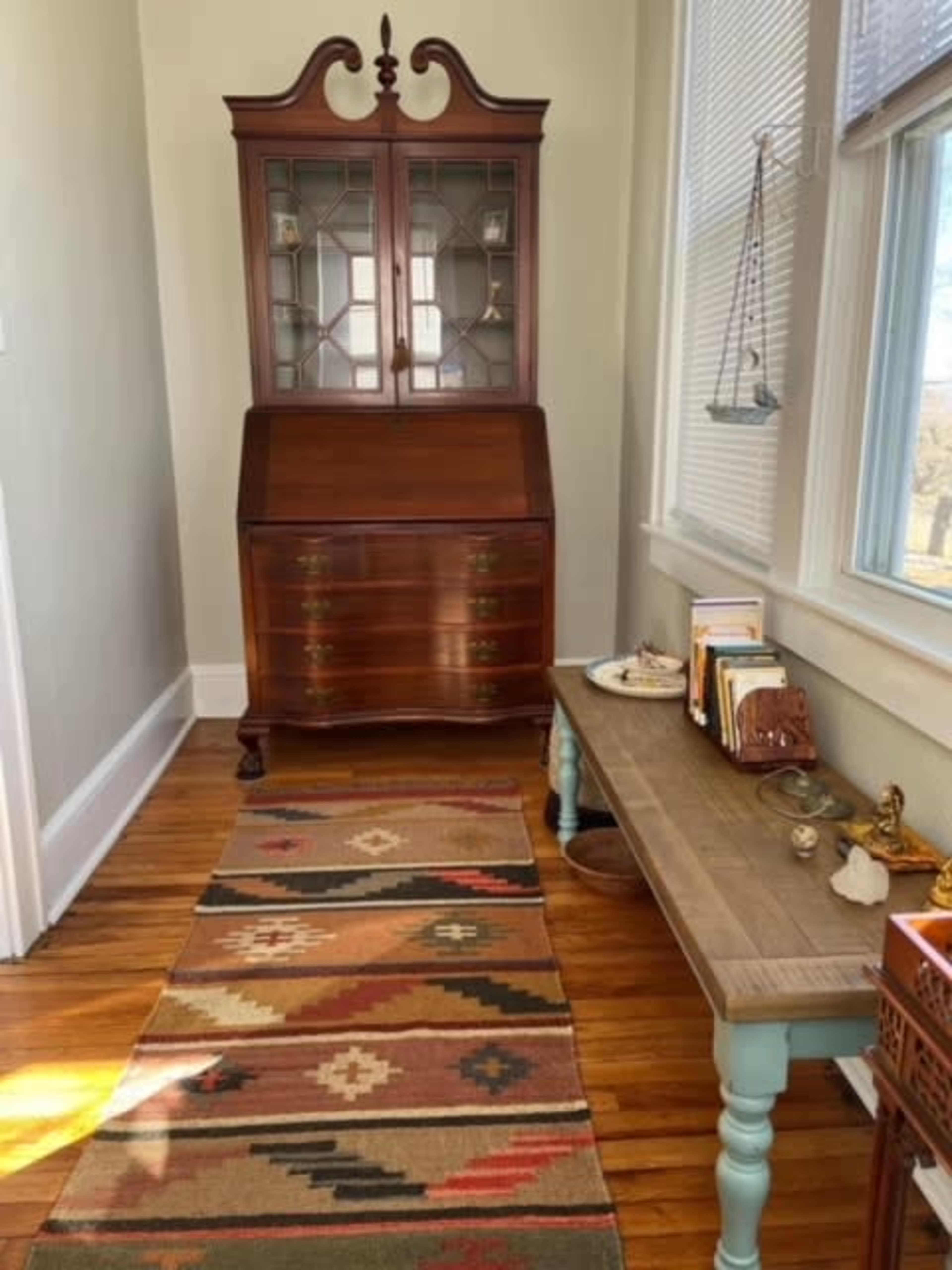 A well-lit hallway featuring a wooden writing desk, a tall glass-fronted cabinet, and a patterned rug on the hardwood floor.