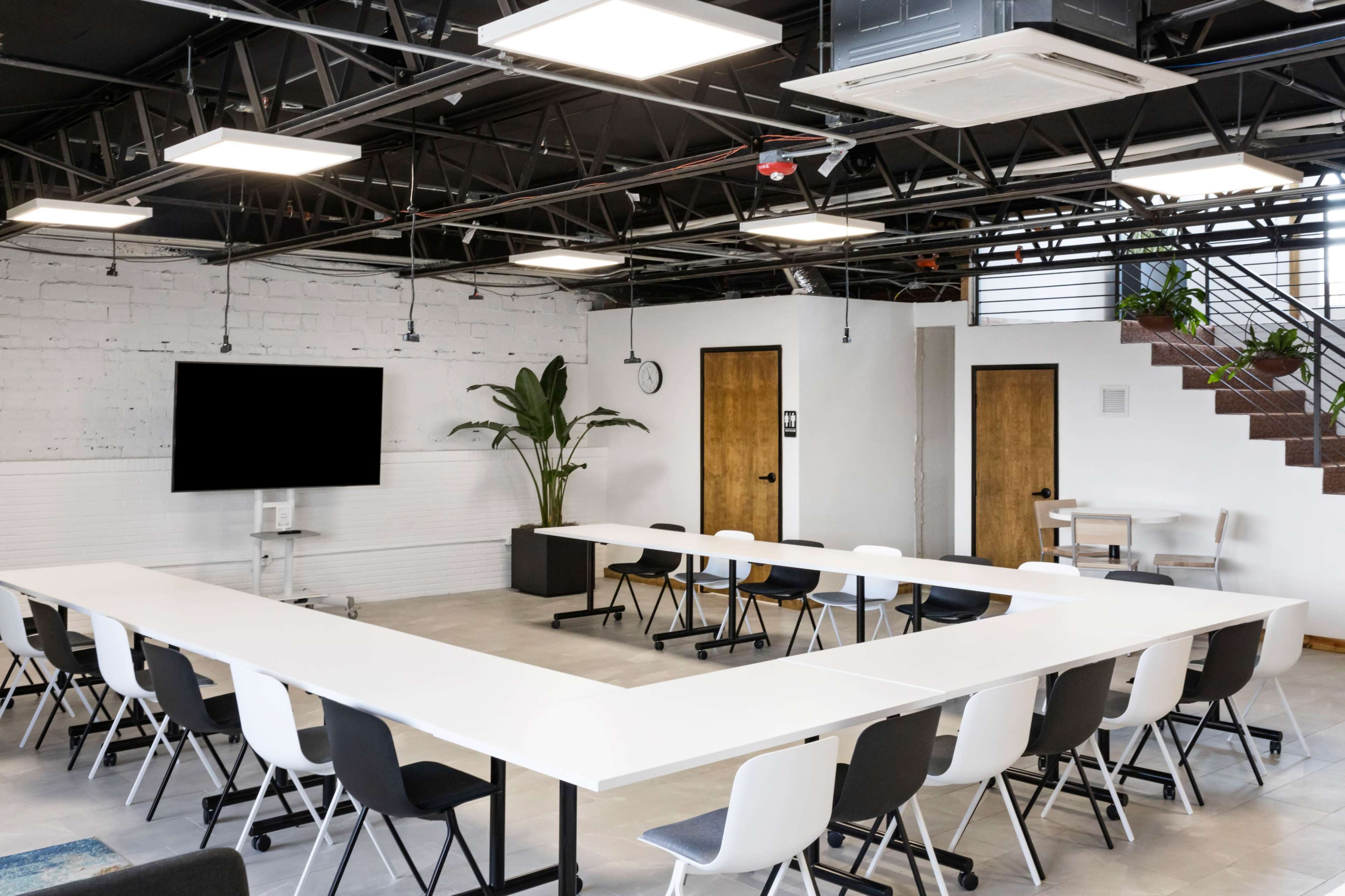 A modern conference room features a large U-shaped table surrounded by chairs, with a television screen on one wall and a staircase in the background.