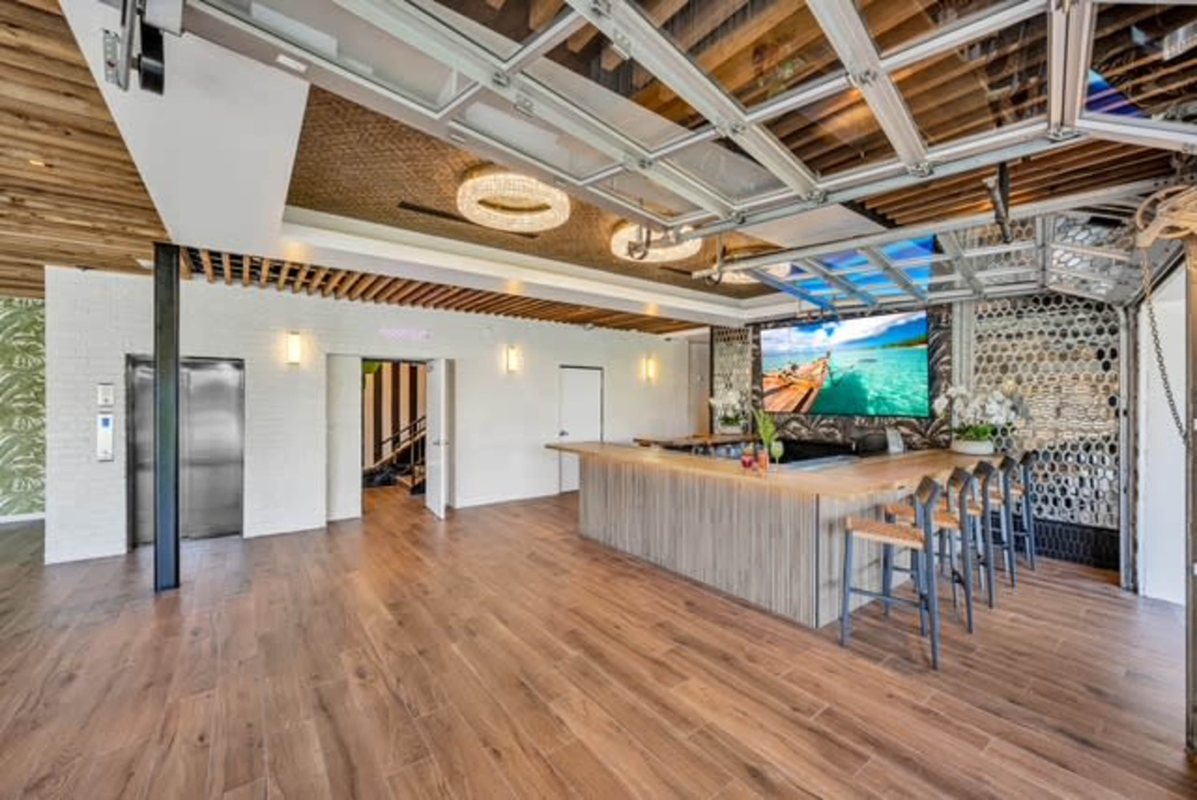 The image shows a modern bar area featuring a wooden countertop, bar stools, a large television displaying a scenic view, and a ceiling with glass panels.