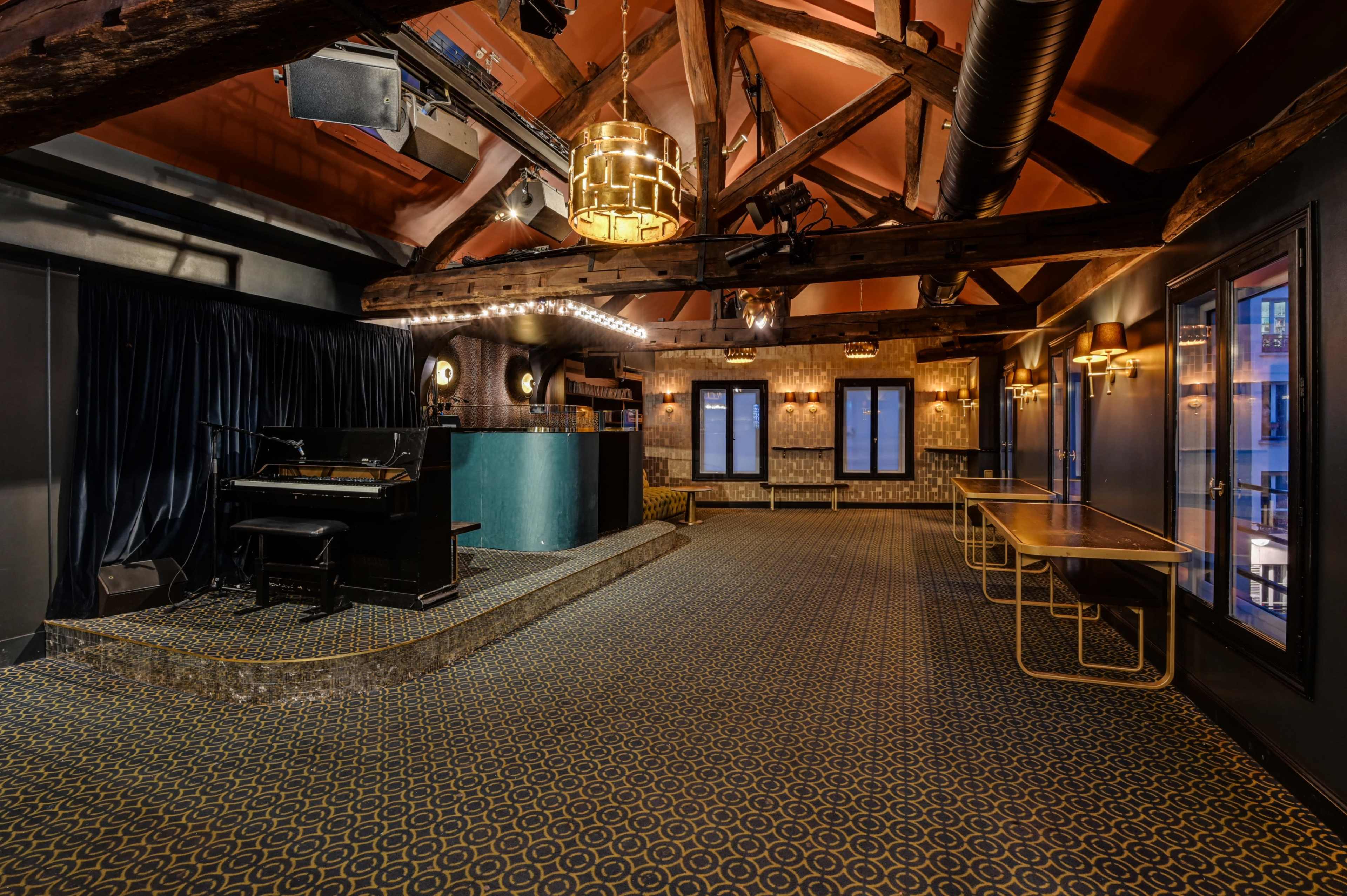 The image depicts a spacious, upscale interior venue featuring a bar area, a piano, exposed wooden beams, and several large windows.
