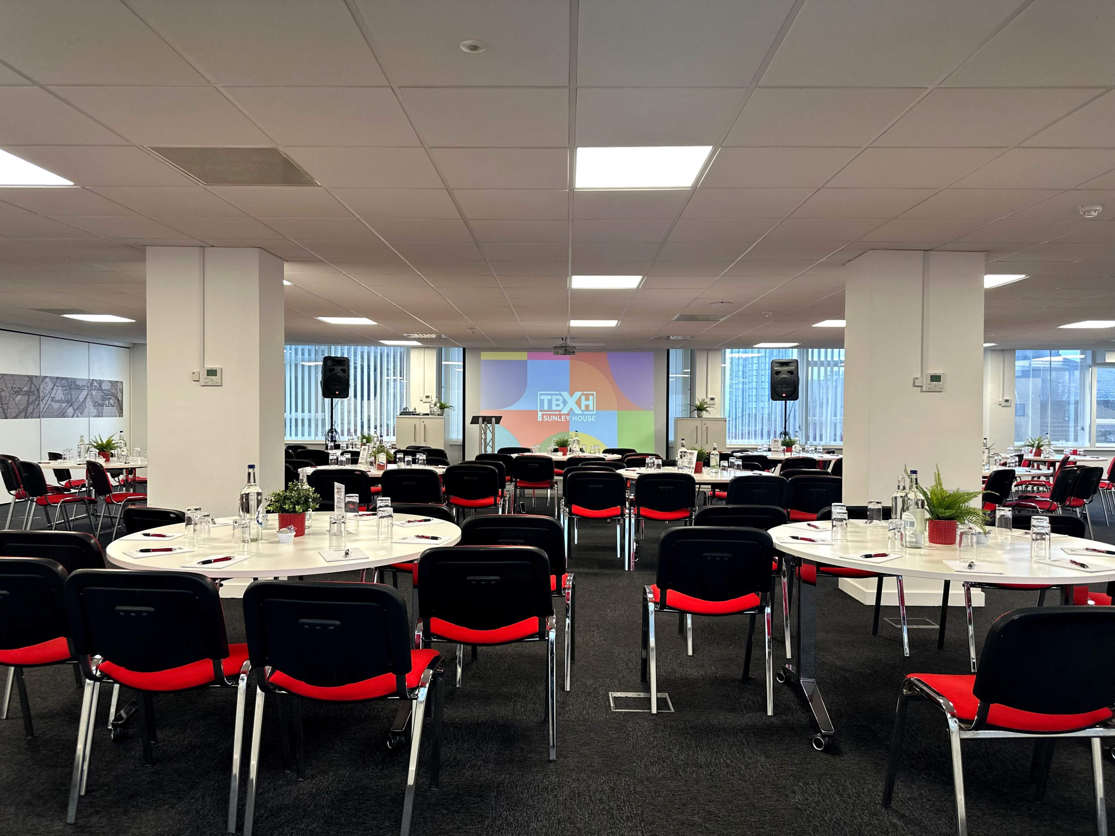 A conference room is set up with round tables arranged for a presentation, featuring chairs, plants, and a screen displaying the event logo.