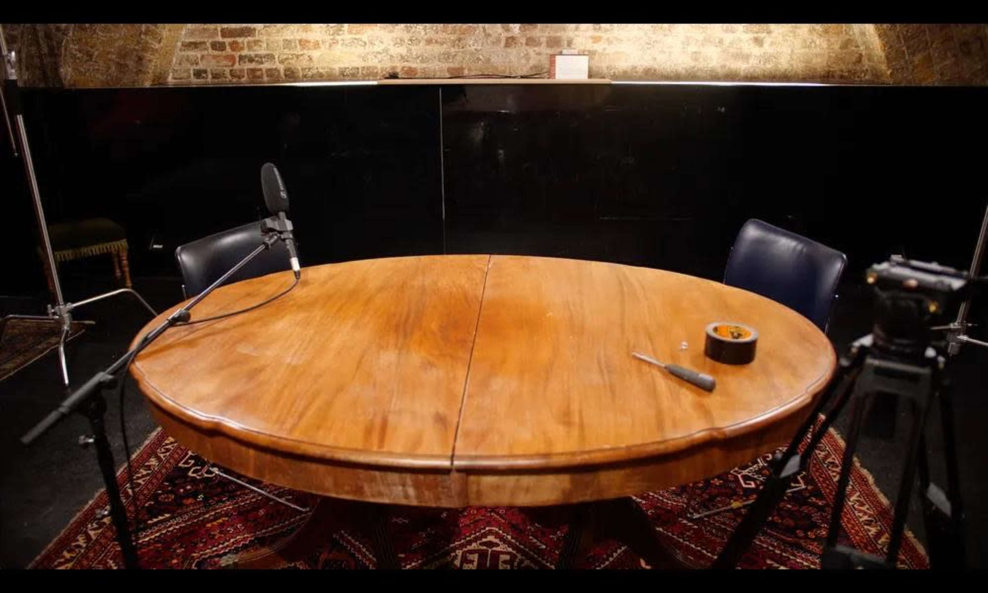 A round wooden table with two chairs and microphones is set up in a dimly lit room with brick walls and a patterned rug.