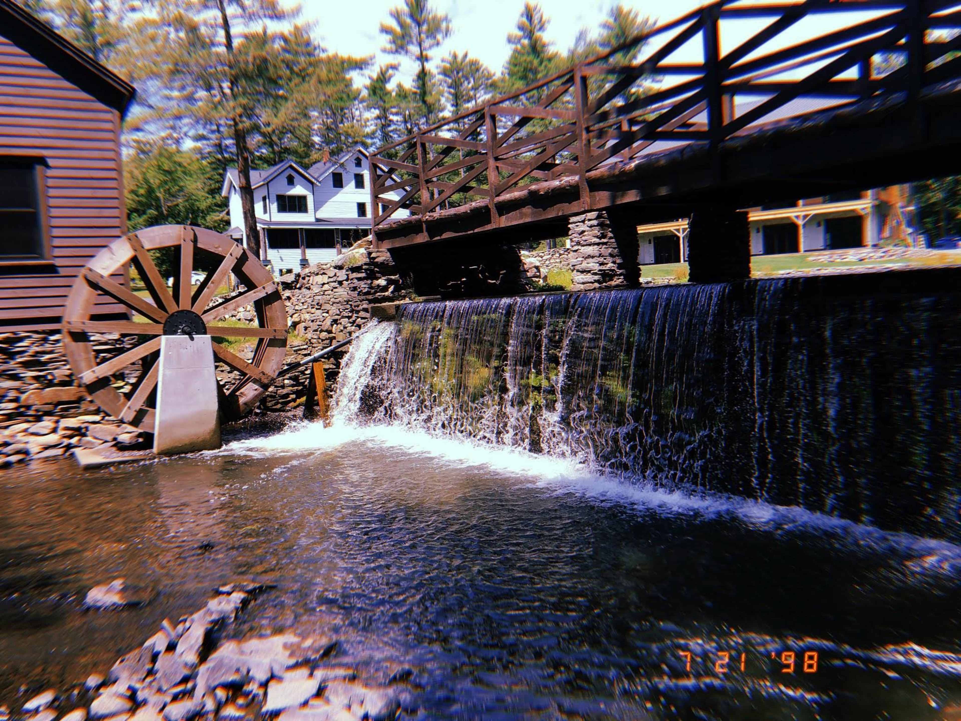 The image shows a wooden water wheel next to a flowing waterfall beneath a bridge, with buildings situated in the background surrounded by trees.
