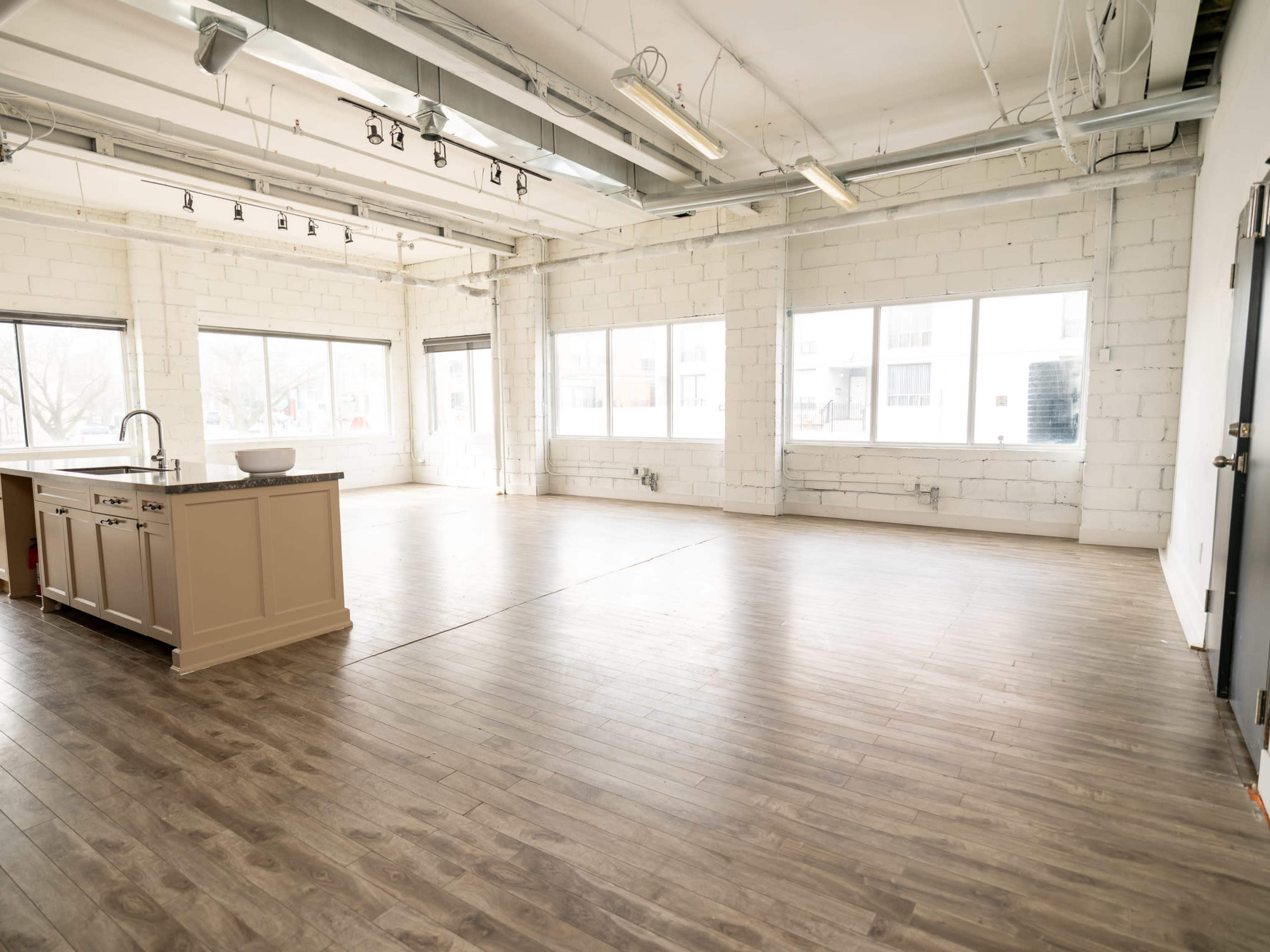 The image shows a large, empty space with wooden flooring, large windows, and a kitchen island at one end.