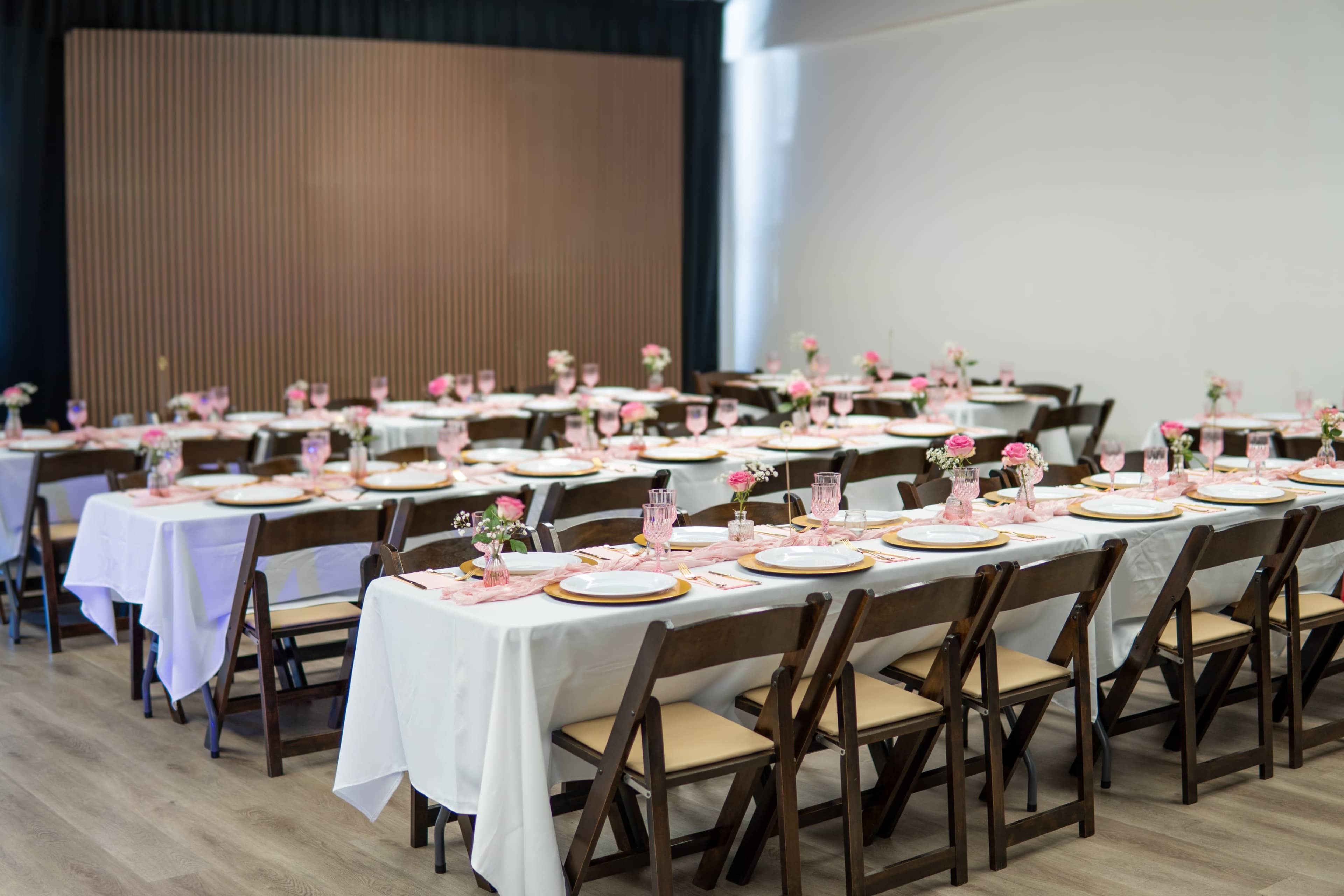 The image shows a banquet setup with white tablecloths, pink floral centerpieces, and plates arranged on long tables in a spacious room.