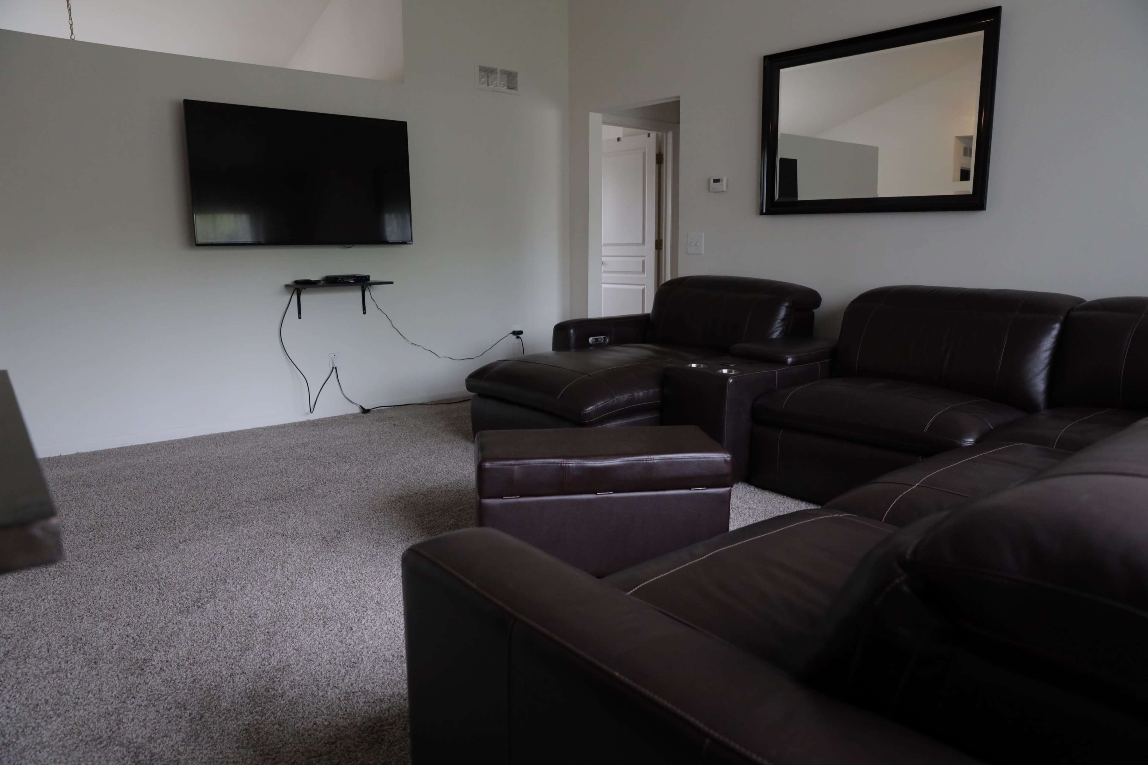 A living room with a brown sectional sofa, a large flat-screen TV mounted on the wall, and a carpeted floor.