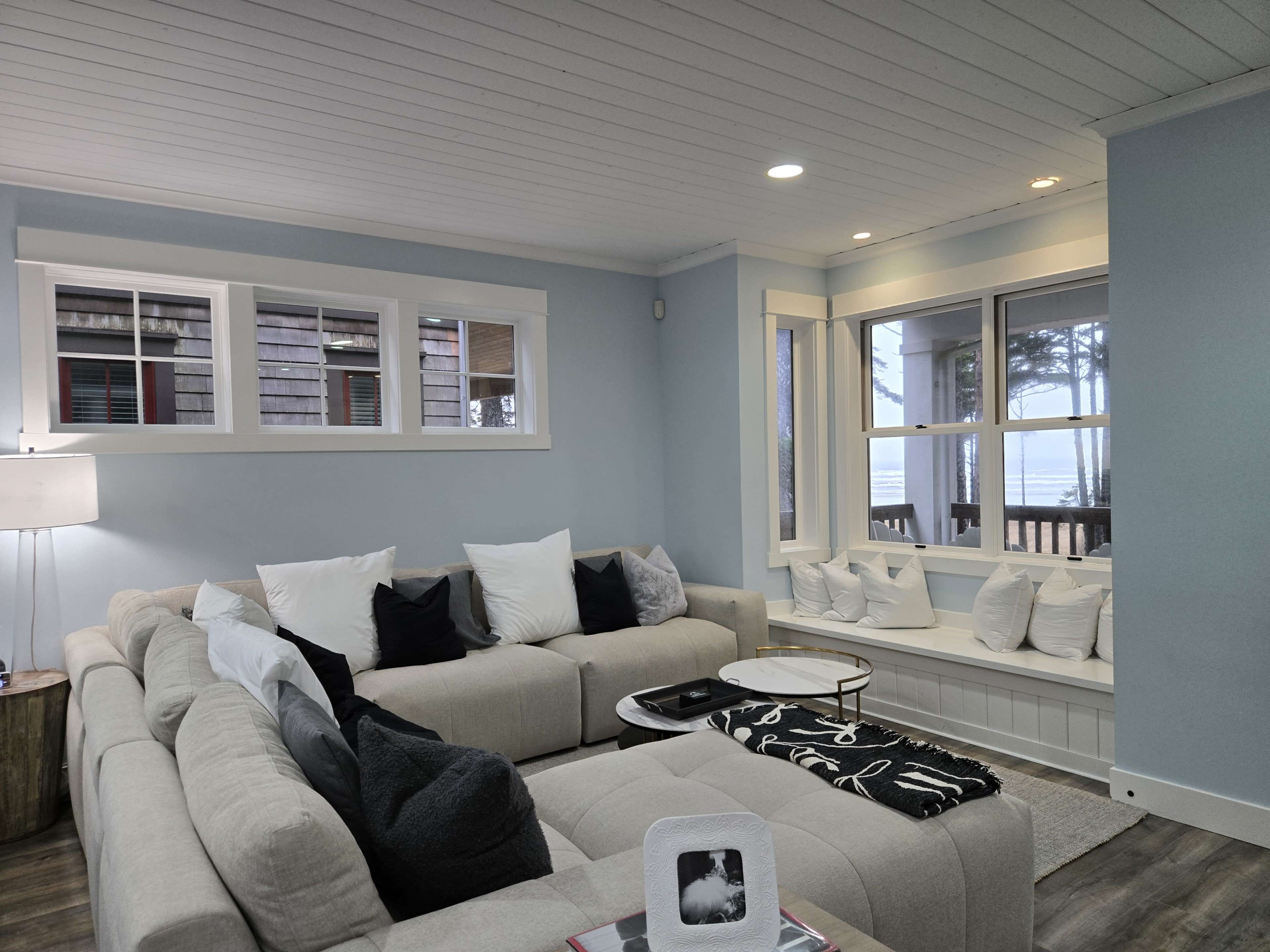The image shows a light blue living room with a large sectional sofa, decorative pillows, and windows that offer a view of the outdoors.