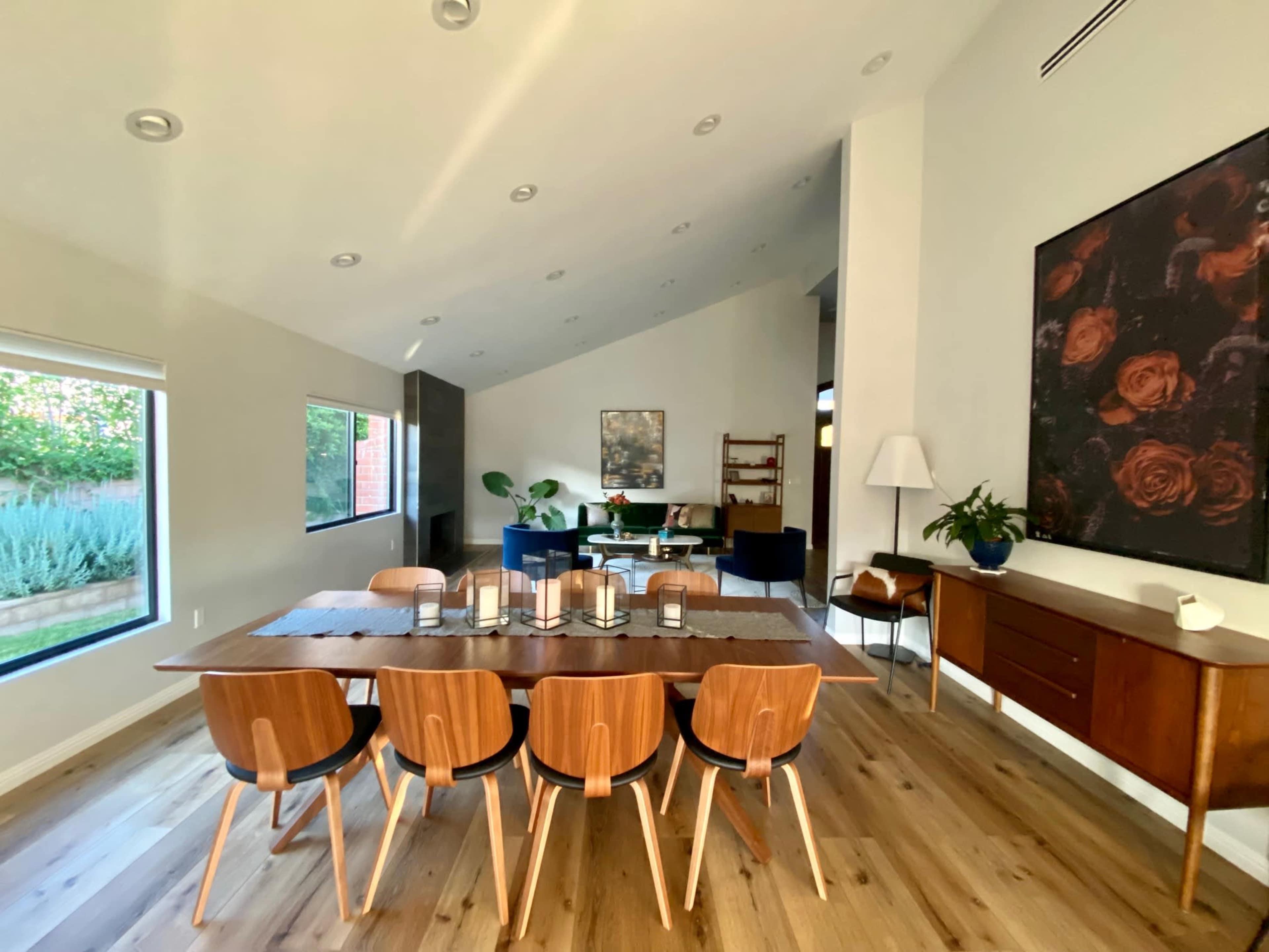 The image shows a modern dining area featuring a wooden table with several chairs, large windows, and a view into a living space with plants and artwork.
