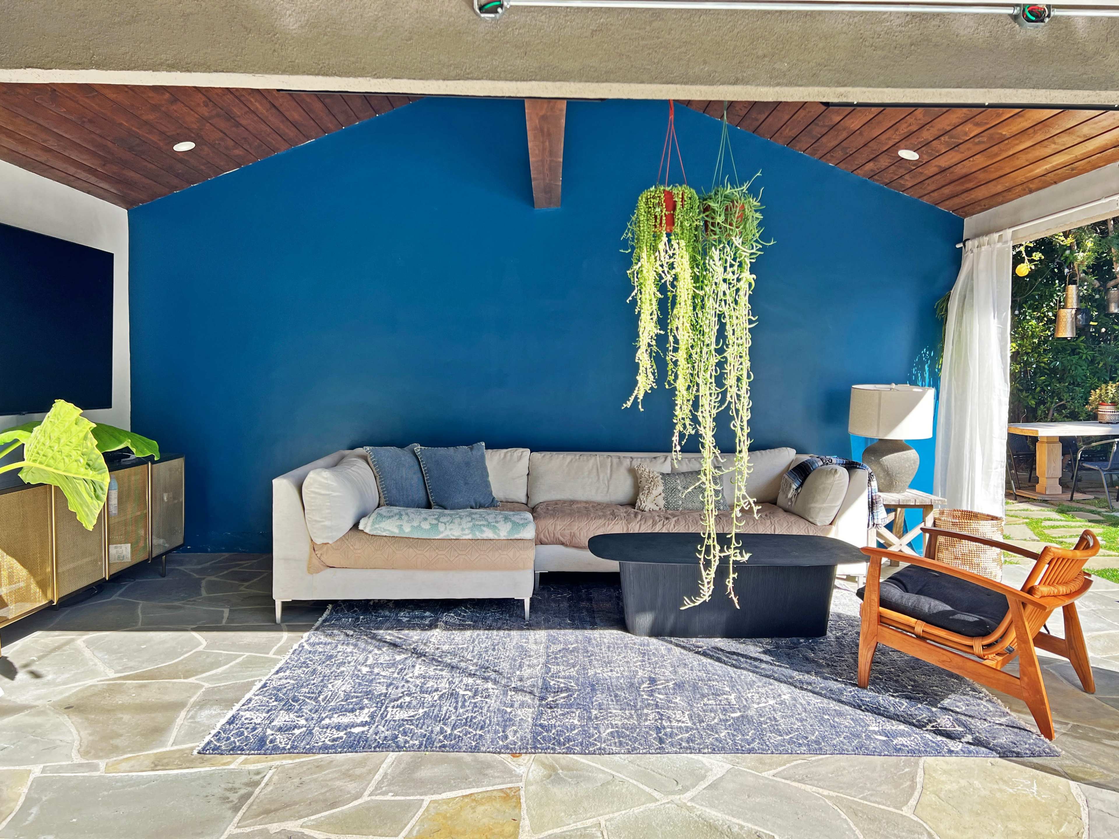 A stylish living area features a beige sectional sofa, a black coffee table, and hanging plants against a blue wall, with large windows overlooking a garden.