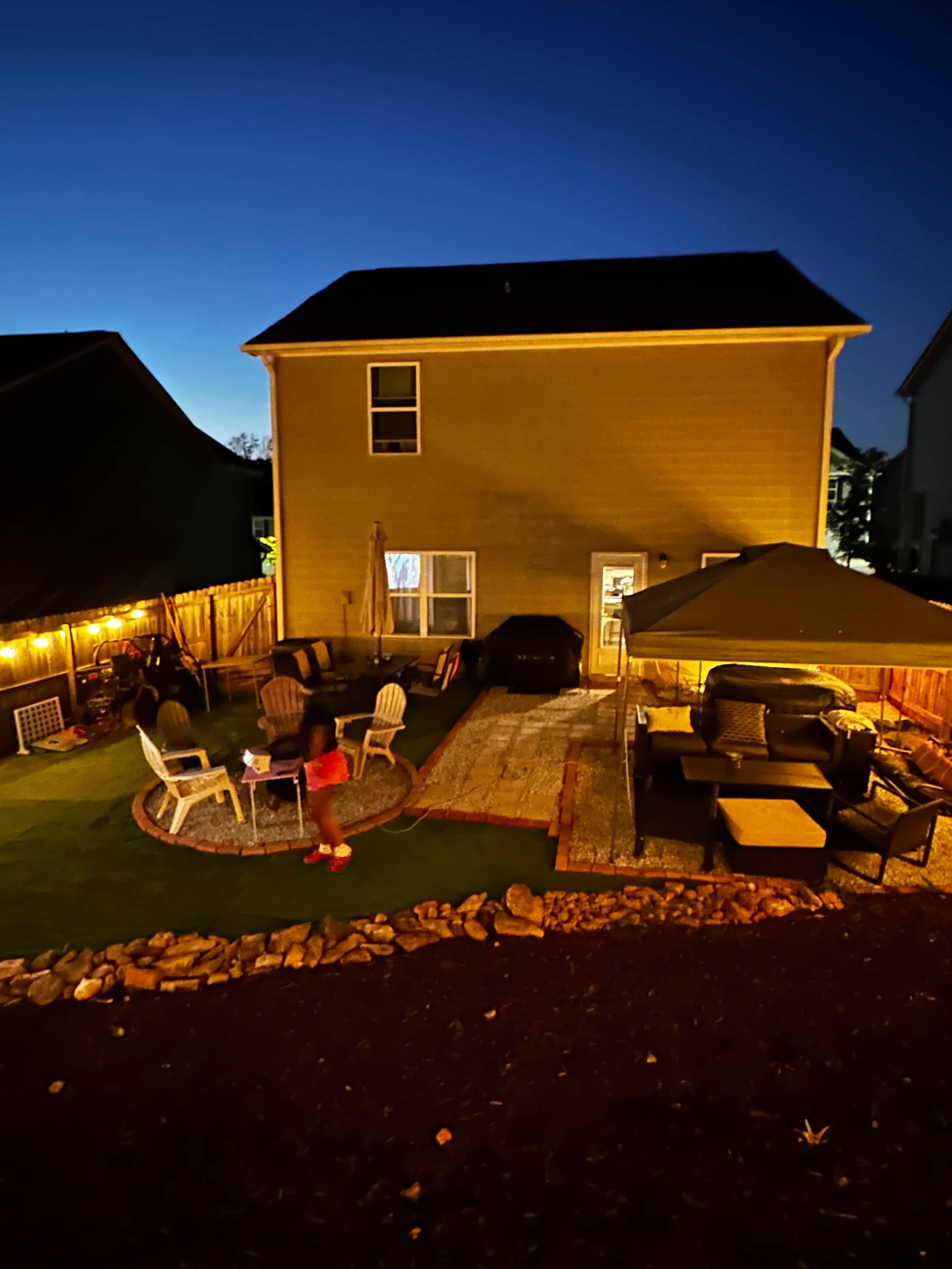 A backyard scene at night features a house with a patio area, outdoor seating, and a large canopy.