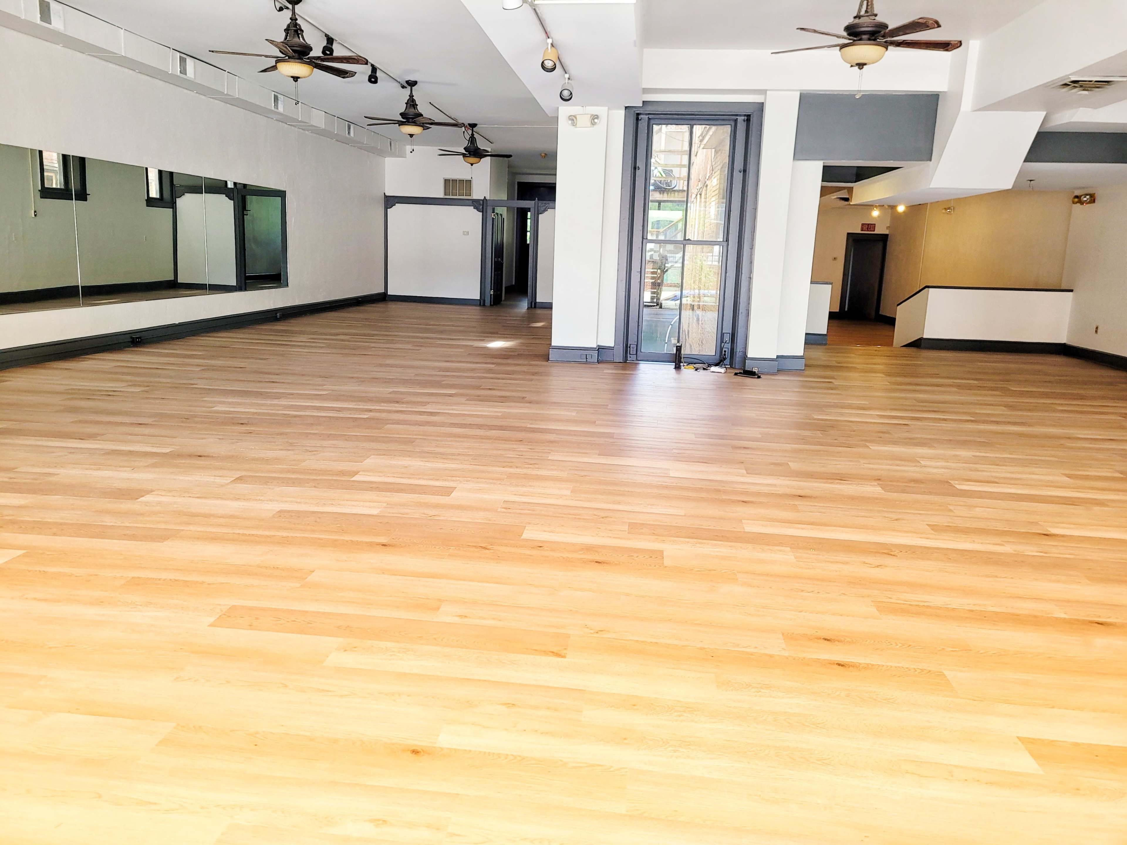 A spacious, empty room with light wooden flooring and large mirrors on one wall.