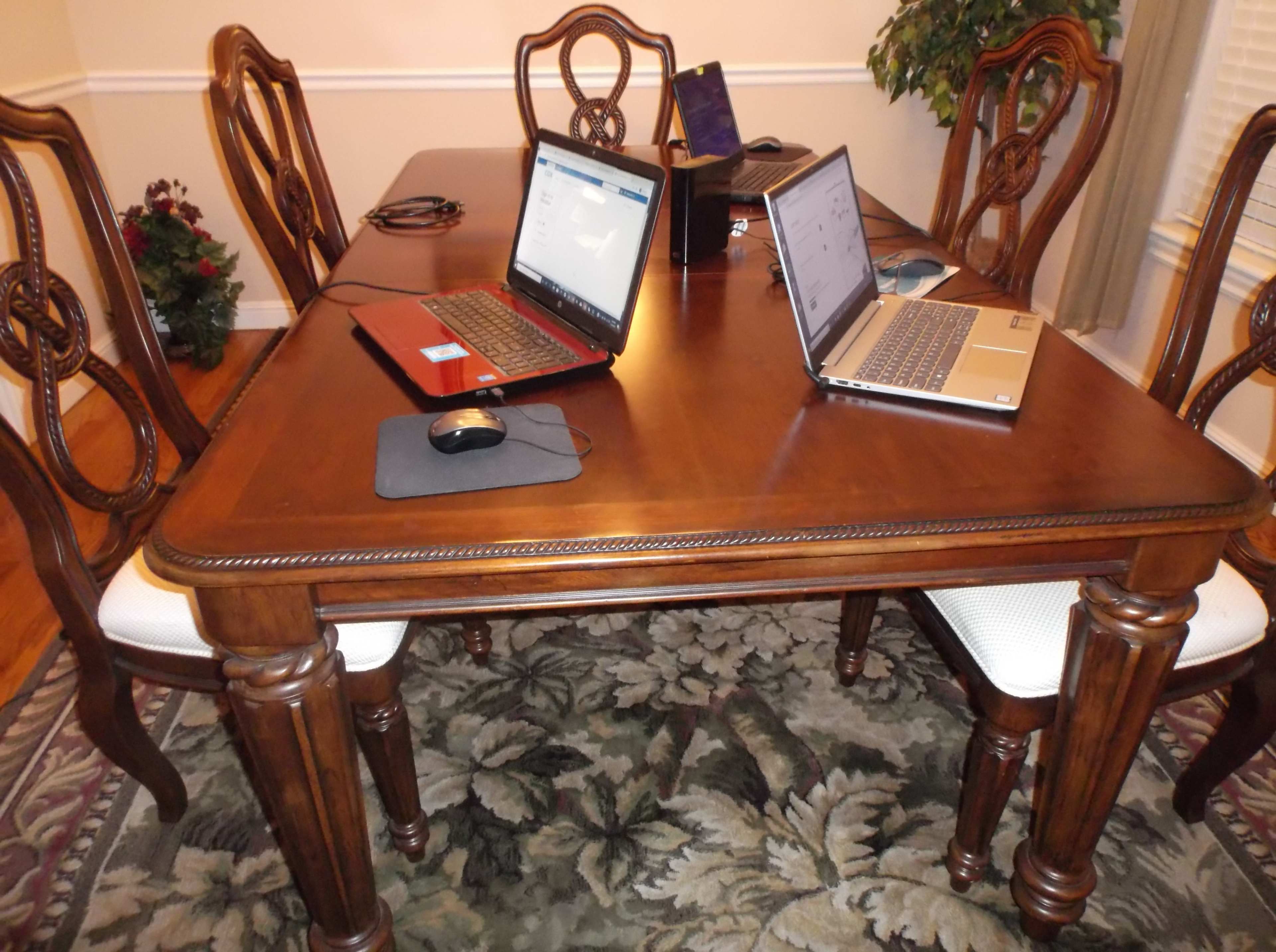 A wooden dining table is set with two laptops and a mouse, surrounded by chairs on a patterned rug.