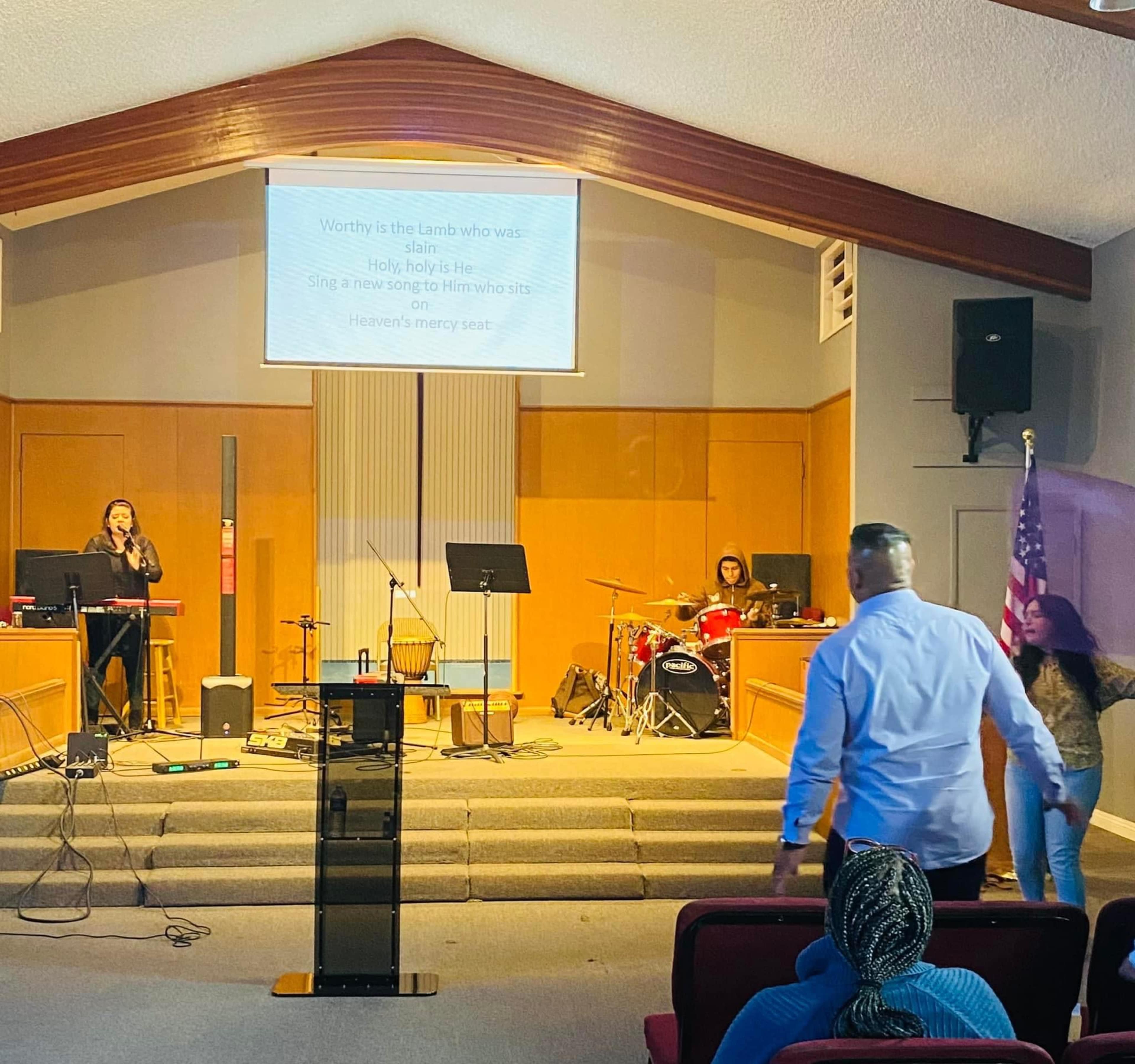 A band performs on stage in a church, while congregants enter the worship area, and lyrics are displayed on a screen.