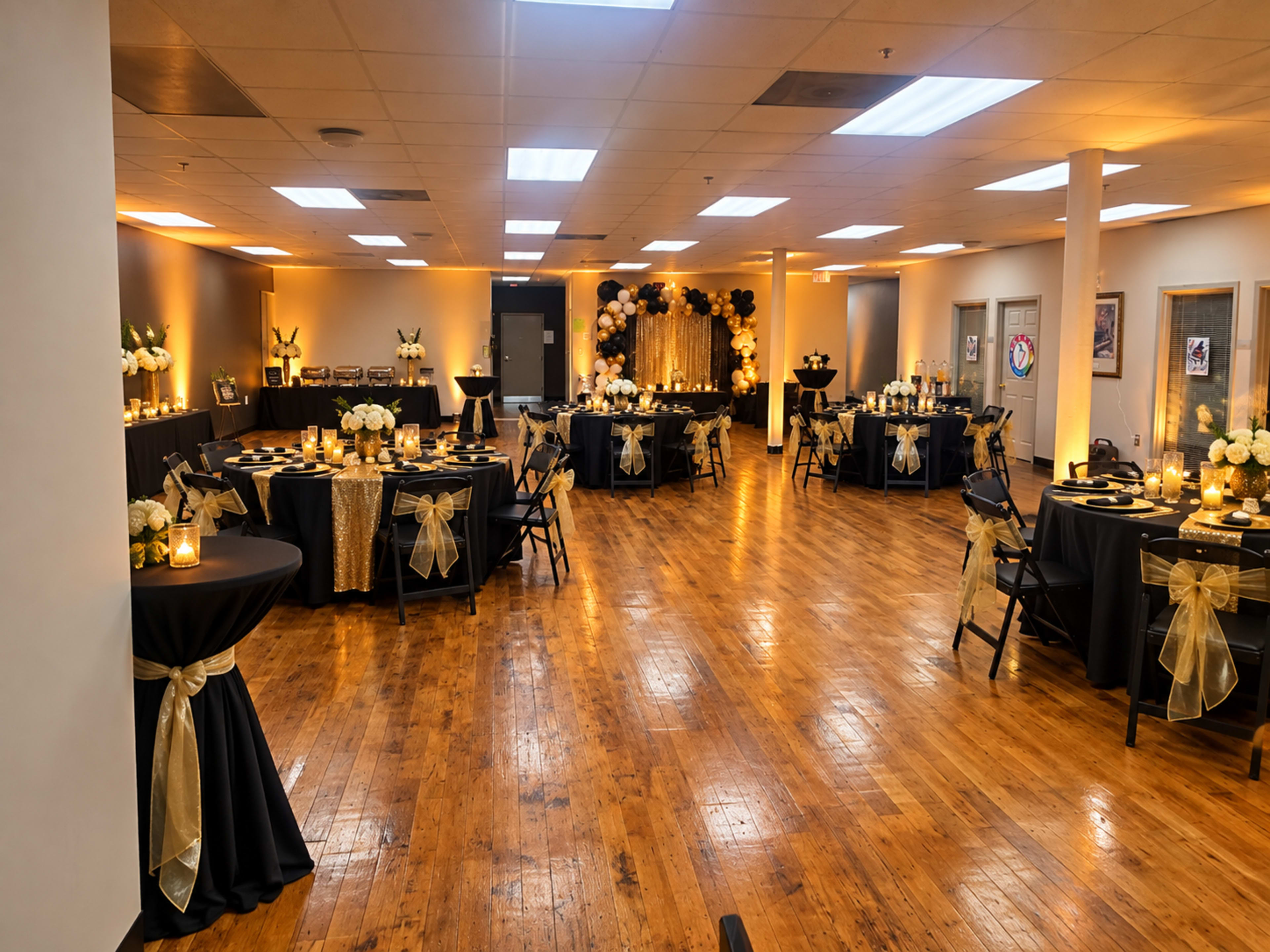 A large event space is set up with black and gold table decorations, featuring round tables covered in black cloth and adorned with floral arrangements and candles.