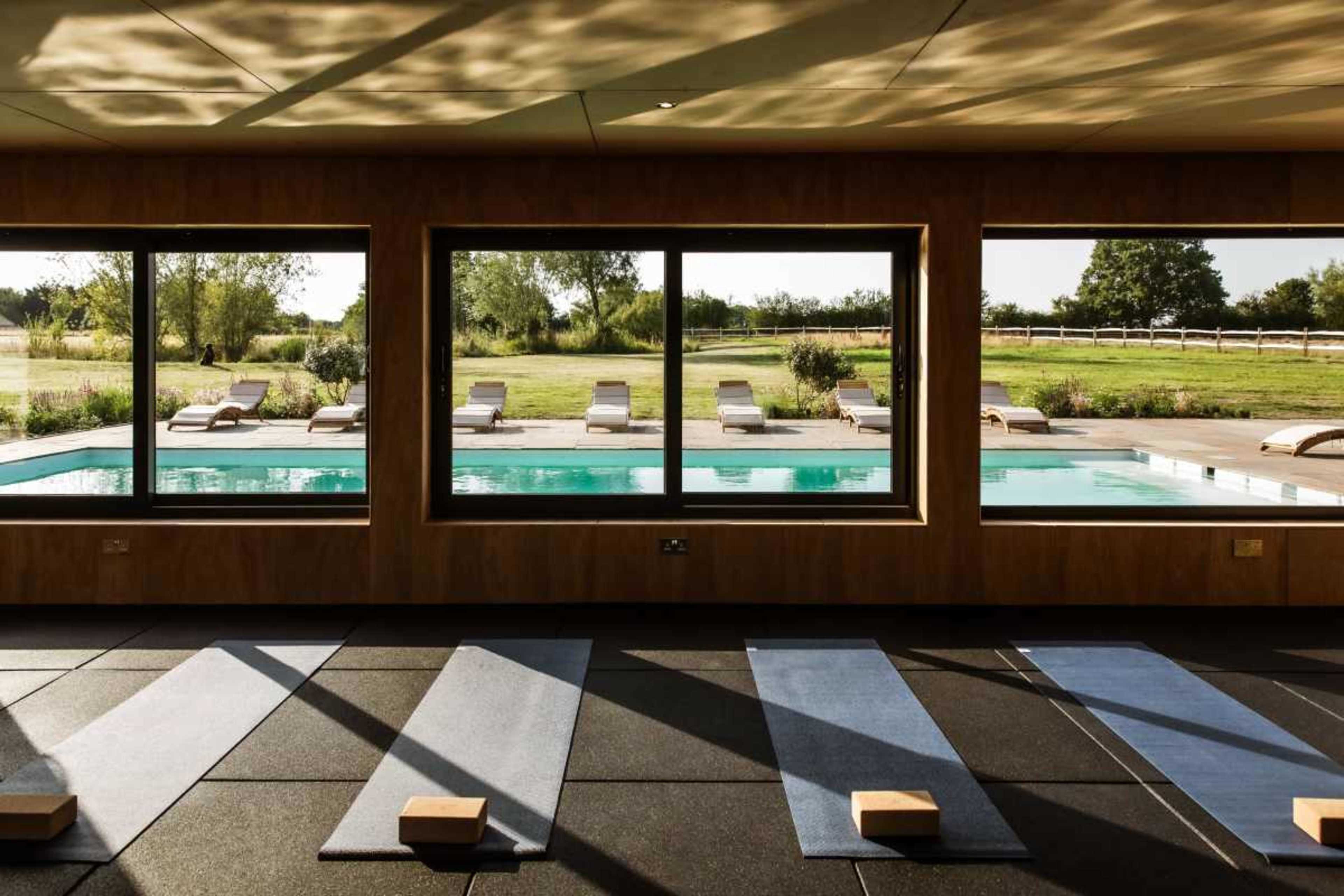 The image shows a yoga studio with mats arranged on the floor, overlooking a serene outdoor pool area surrounded by greenery.