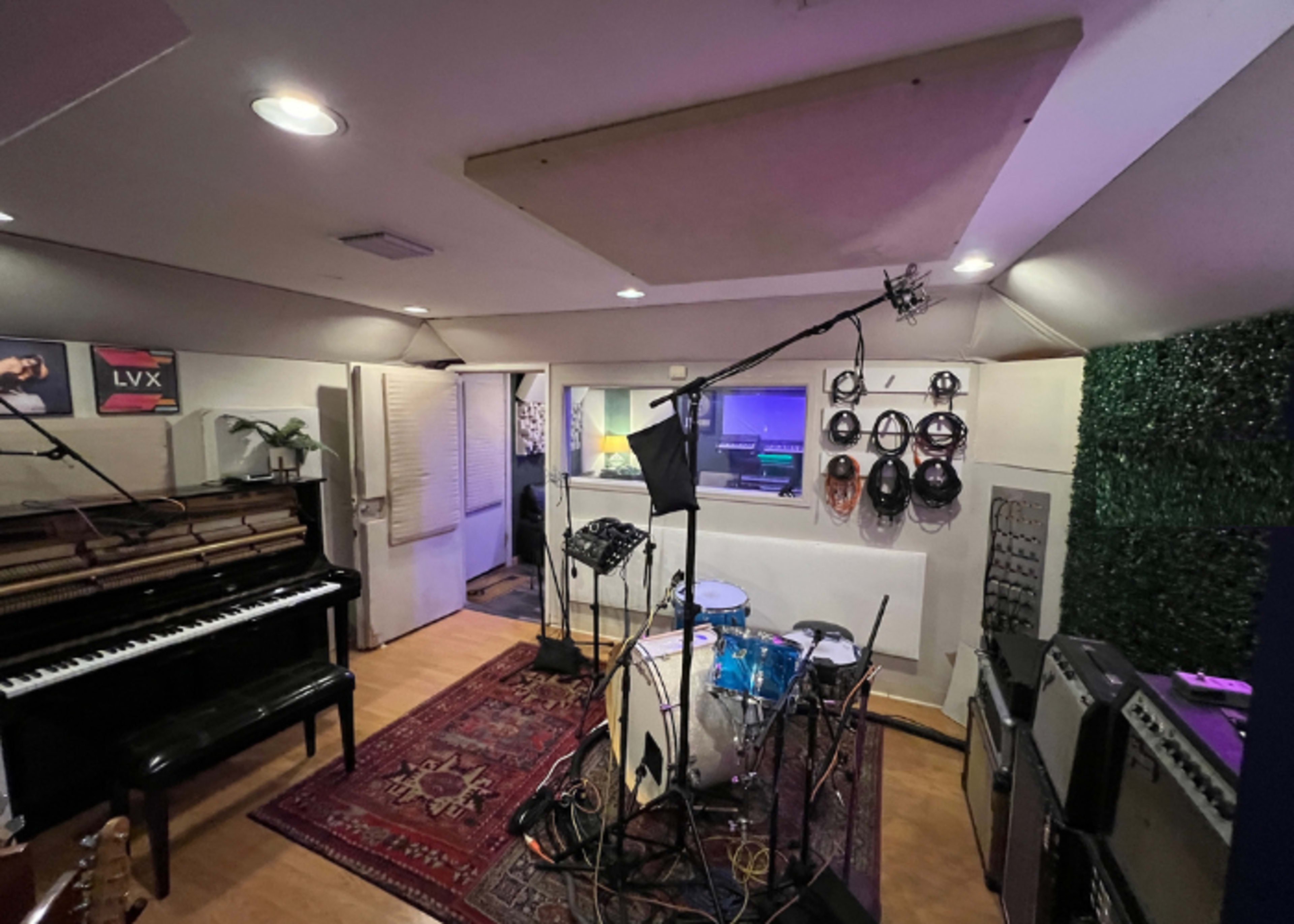 The image shows a music studio equipped with a drum set, a piano, and various musical equipment arranged around a patterned rug.